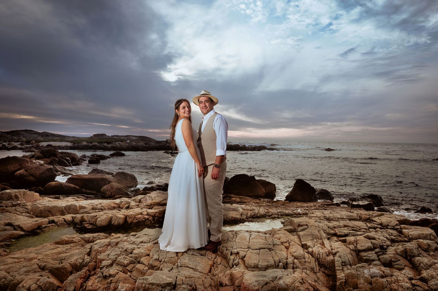Bride and groom posing romantically on rocky coastal shoreline at dramatic sunset.