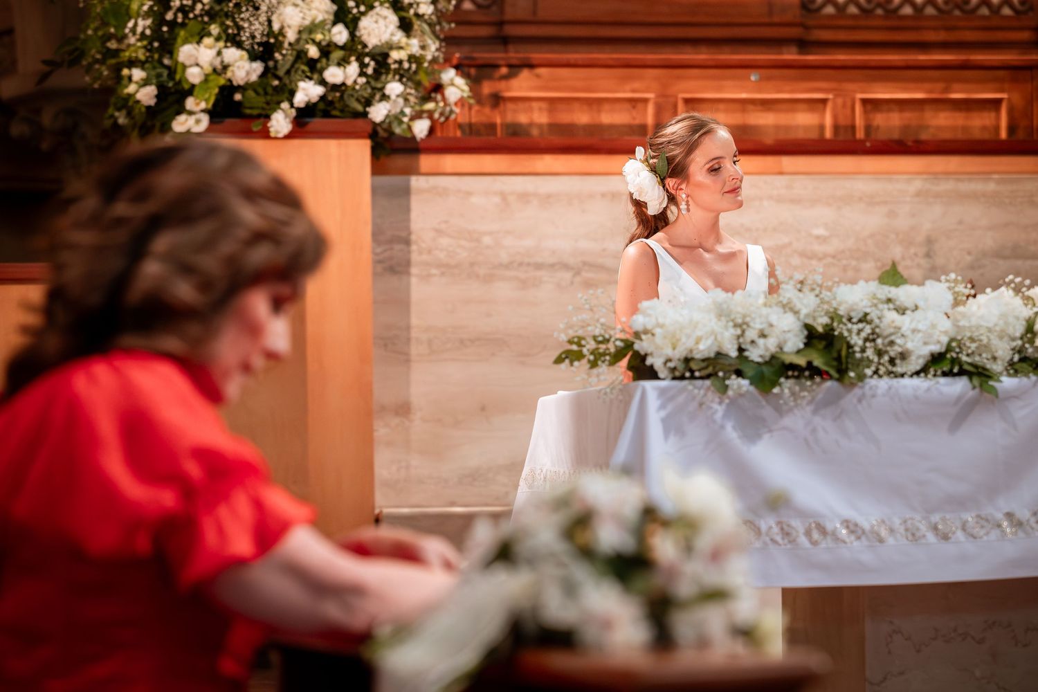 Bride in white dress with floral headpiece sits at altar while guest in red dress watches ceremony.