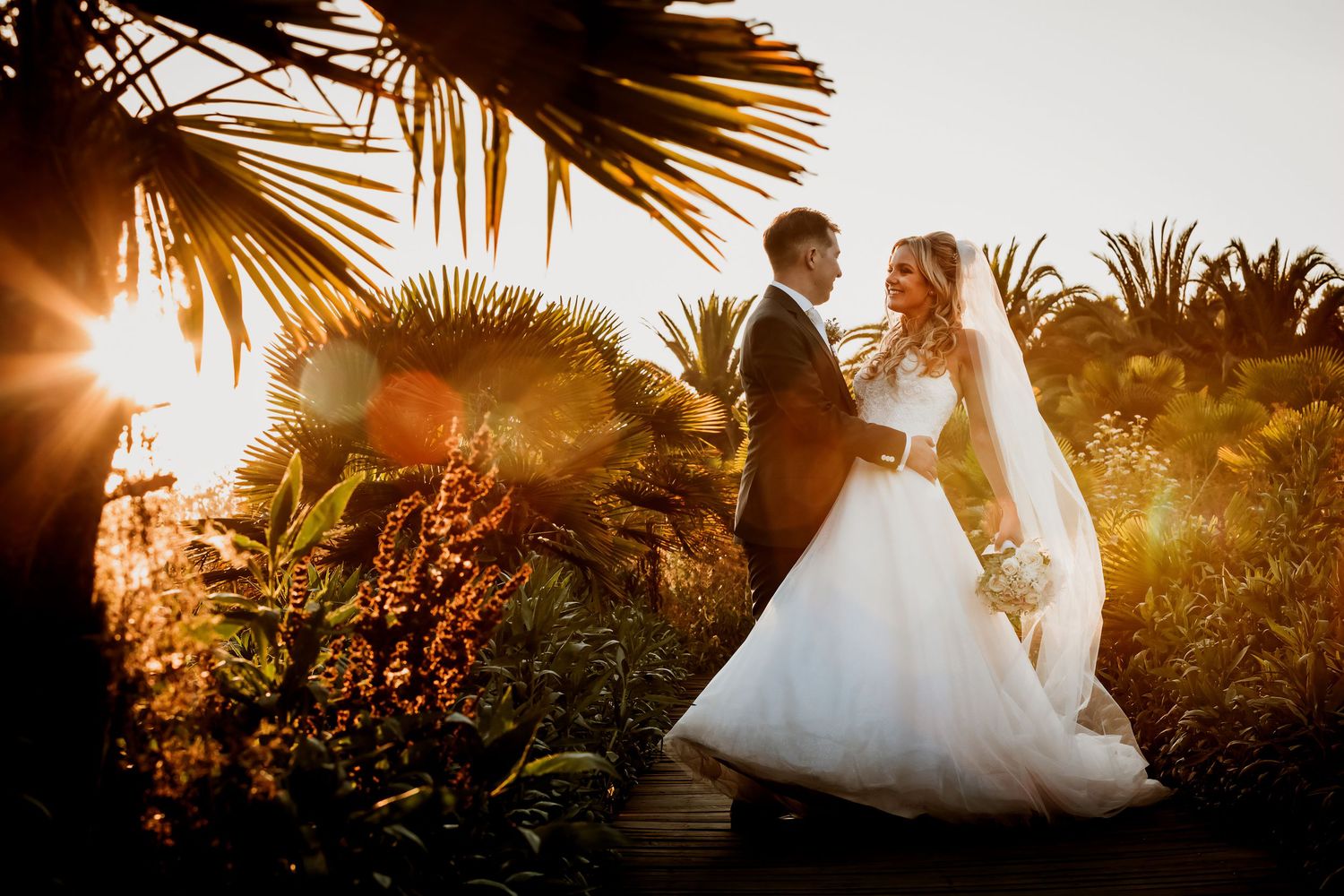 Romantic bride and groom sharing a moment in a sun-drenched tropical garden at sunset.
