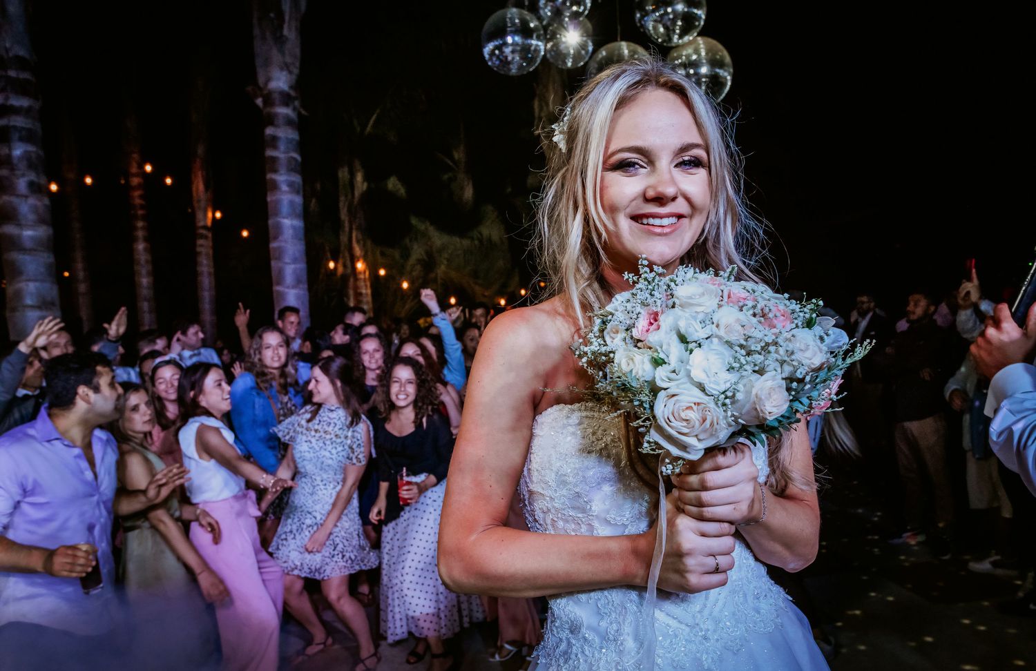 Beautiful blonde bride smiling and holding white and pink bouquet at outdoor evening wedding reception.