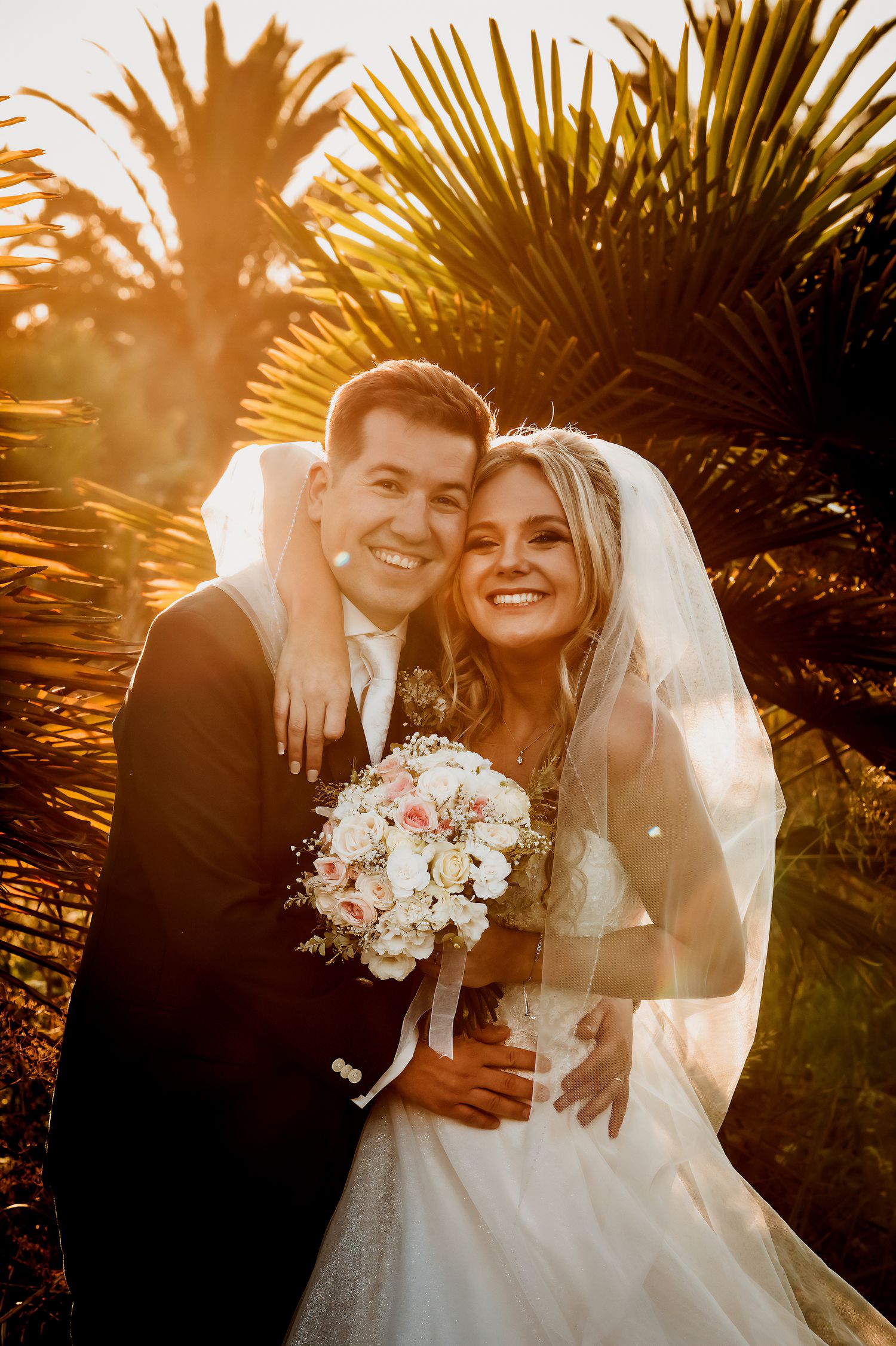 Happy newlyweds embracing in warm golden sunlight with tropical palm trees in the background.