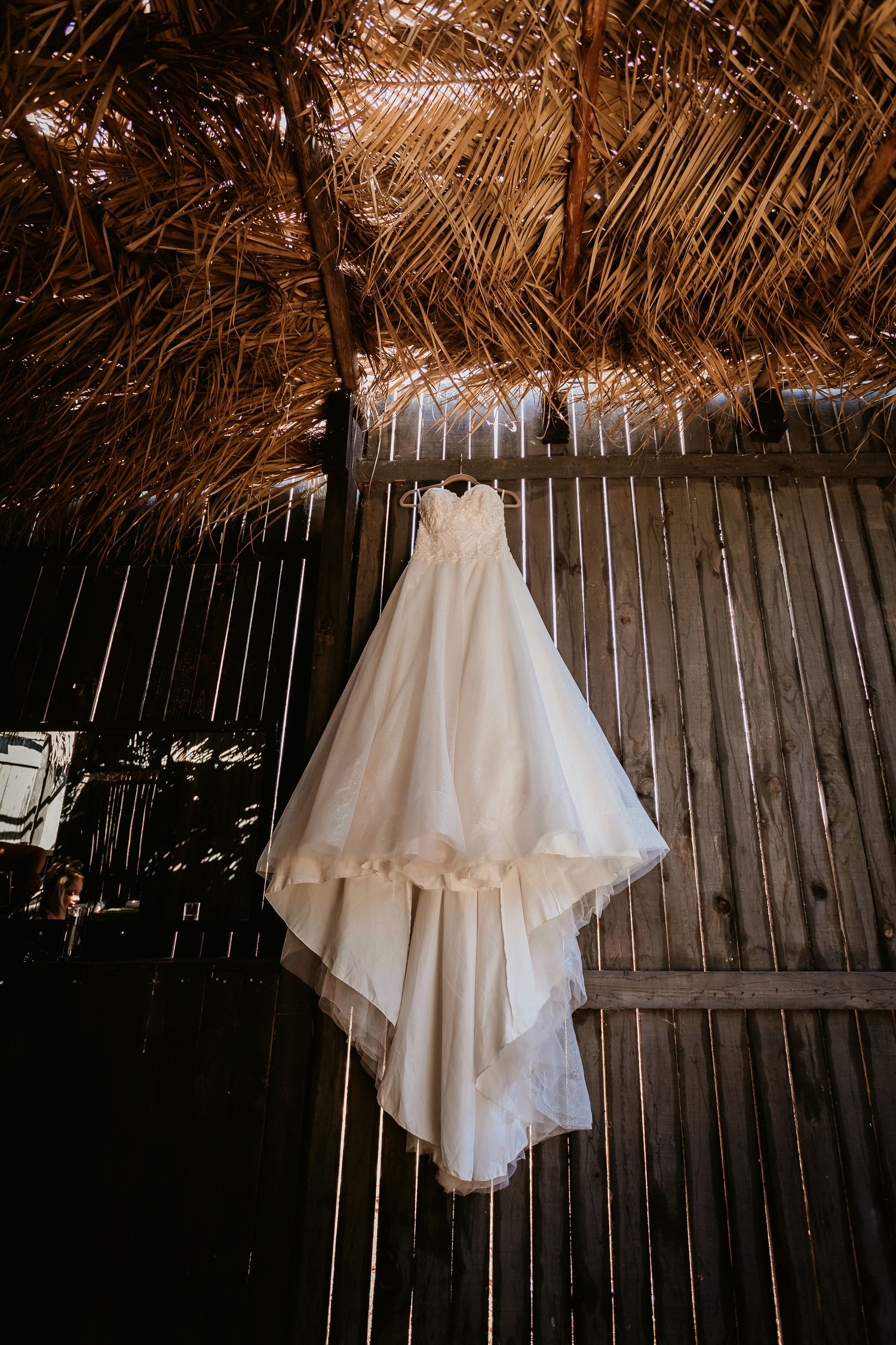 An elegant strapless white wedding gown hangs against a rustic wooden wall beneath a thatched roof.