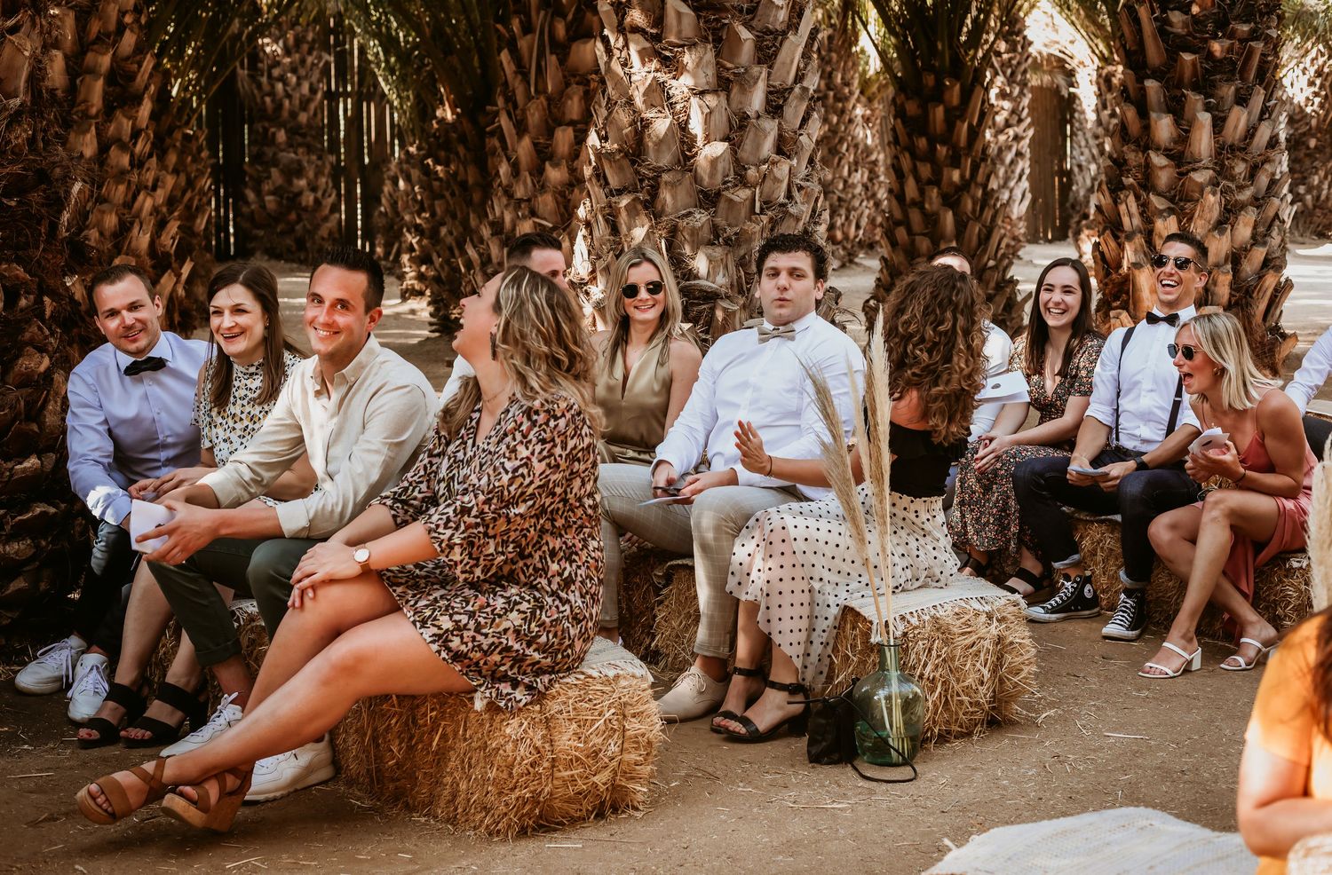 Wedding guests seated on hay bales under palm trees at rustic outdoor ceremony venue.