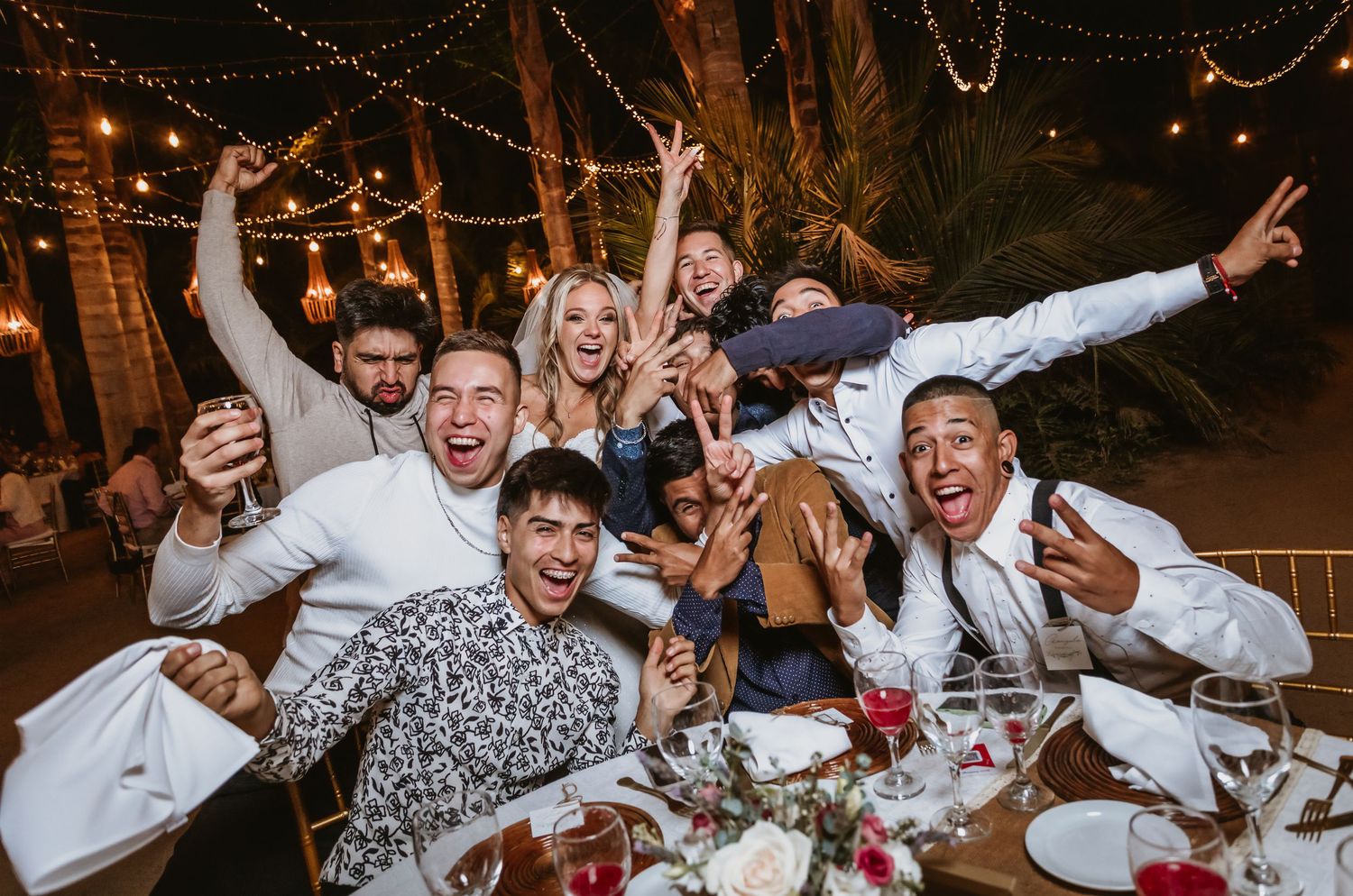 Excited wedding guests celebrate at outdoor reception table under string lights at night.