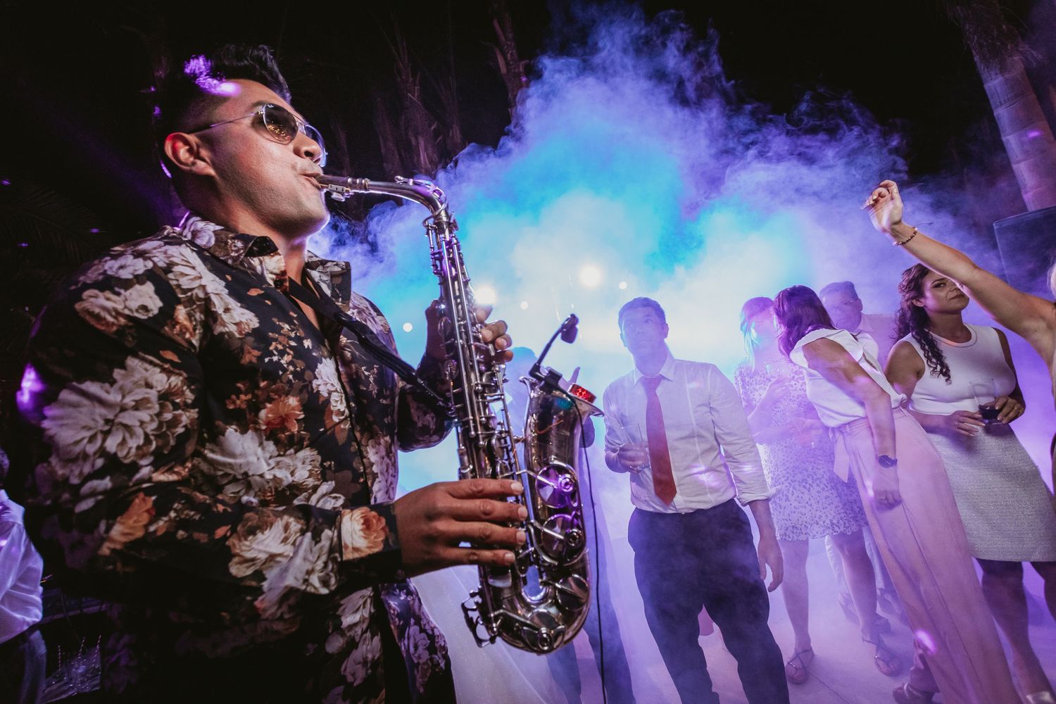 Saxophonist performs in dramatic smoke and lighting on crowded wedding reception dance floor.