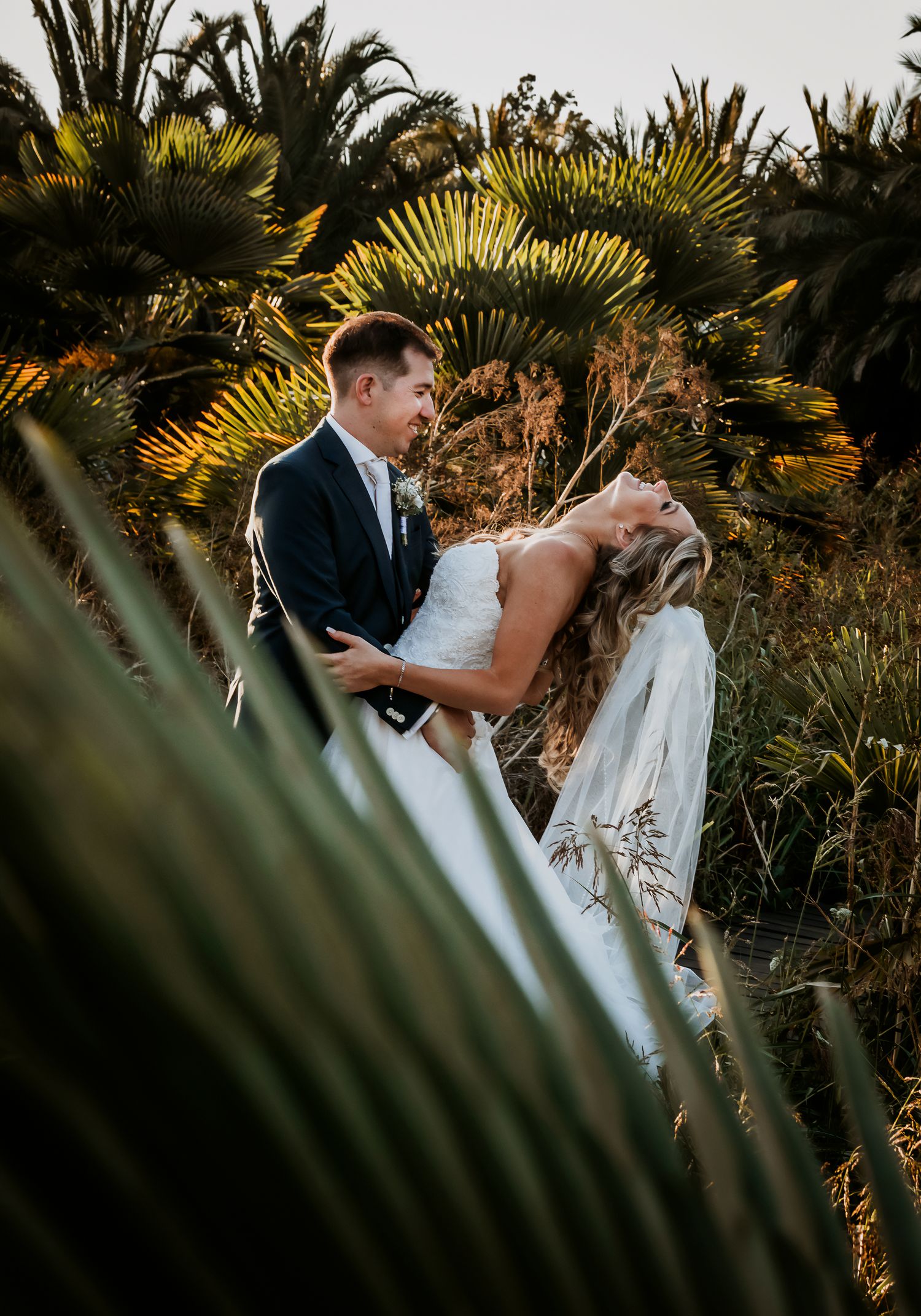 Groom lifts laughing bride among lush tropical palms captured through foreground foliage at sunset.