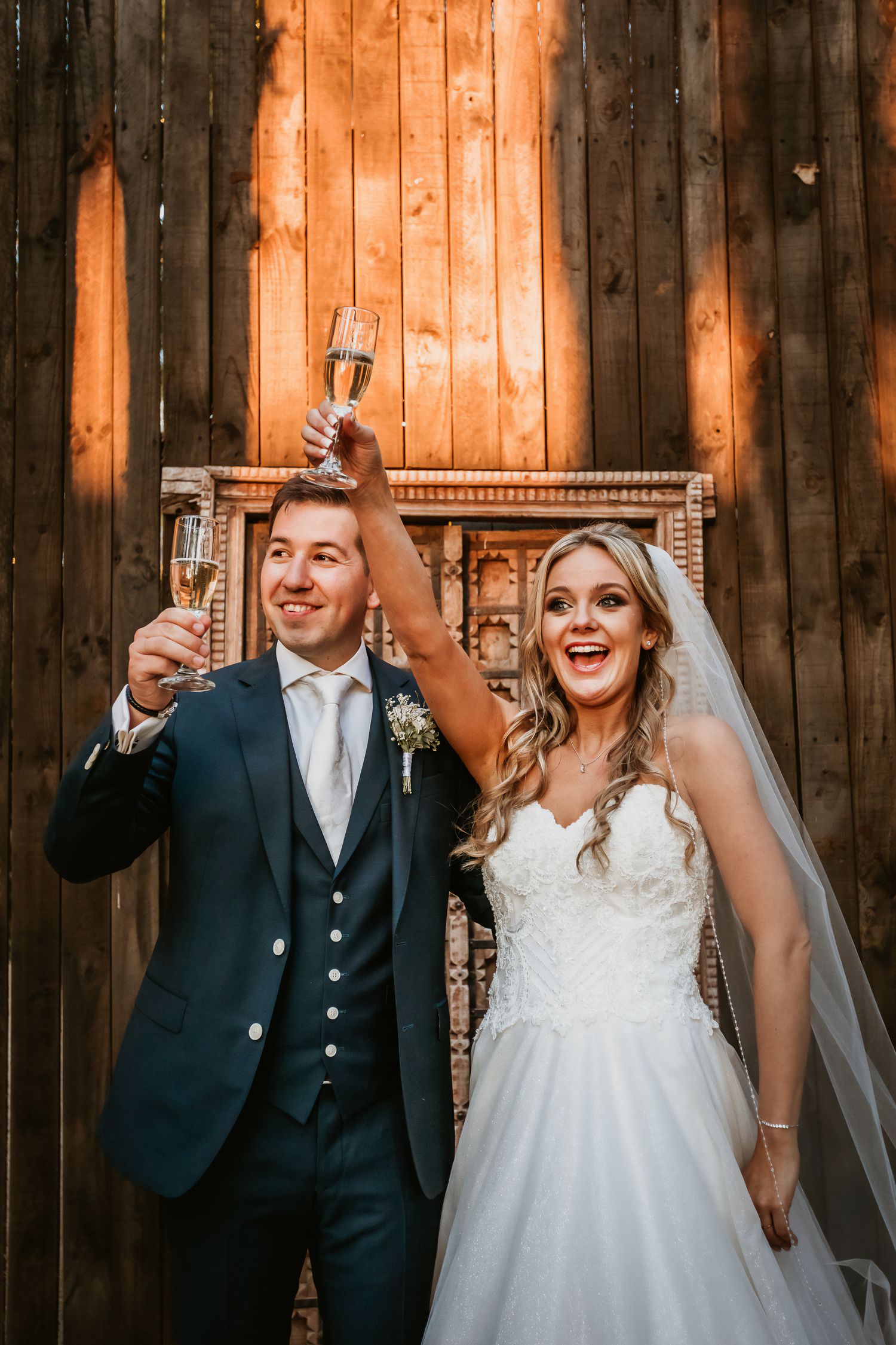 Excited bride and groom raise champagne glasses in celebration against rustic wooden barn backdrop.