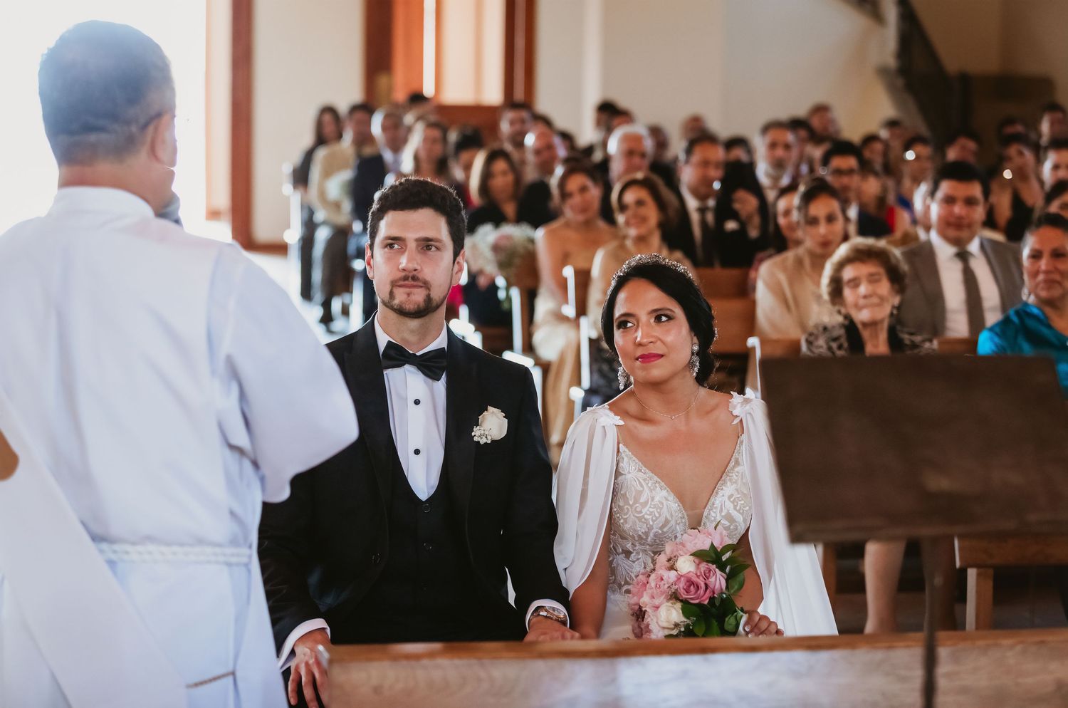 Bride and groom standing before officiant during church wedding ceremony with guests.