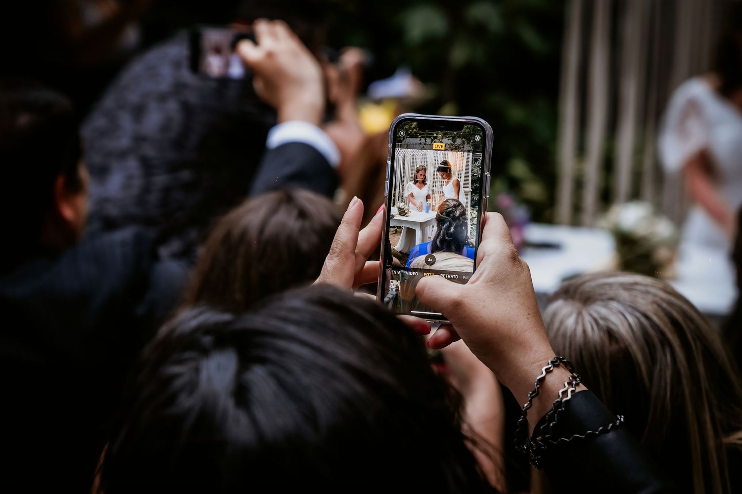 Wedding guest photographs ceremony moment on smartphone as guests gather in garden for outdoor wedding celebration.