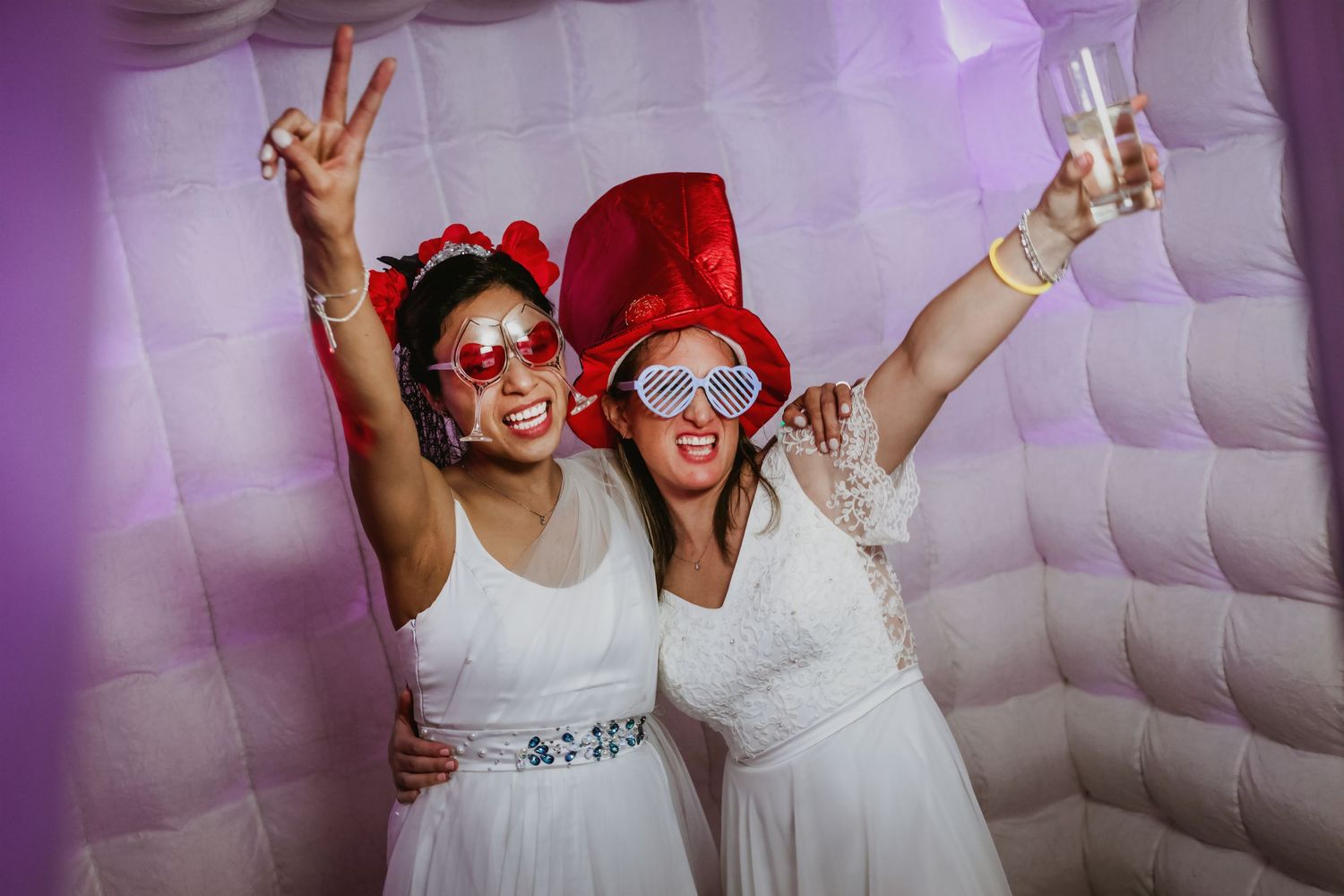 Two brides in white dresses wear oversized sunglasses and fun hats, celebrating in a photo booth together.