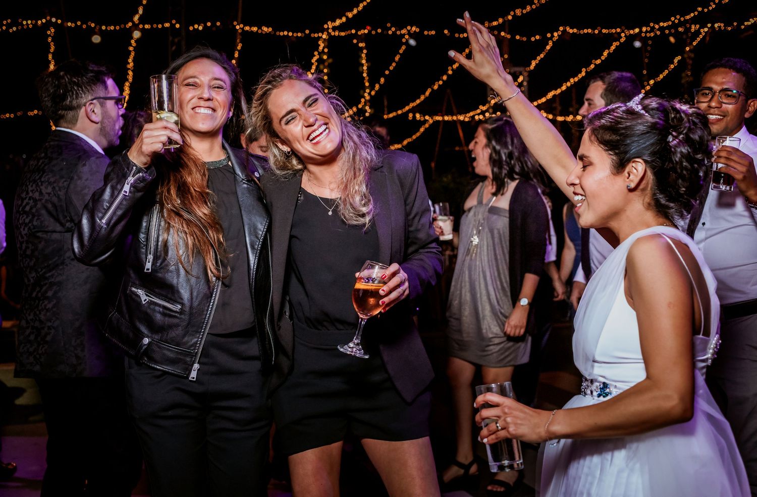 Three women laugh and dance with champagne glasses at a festive wedding reception party.
