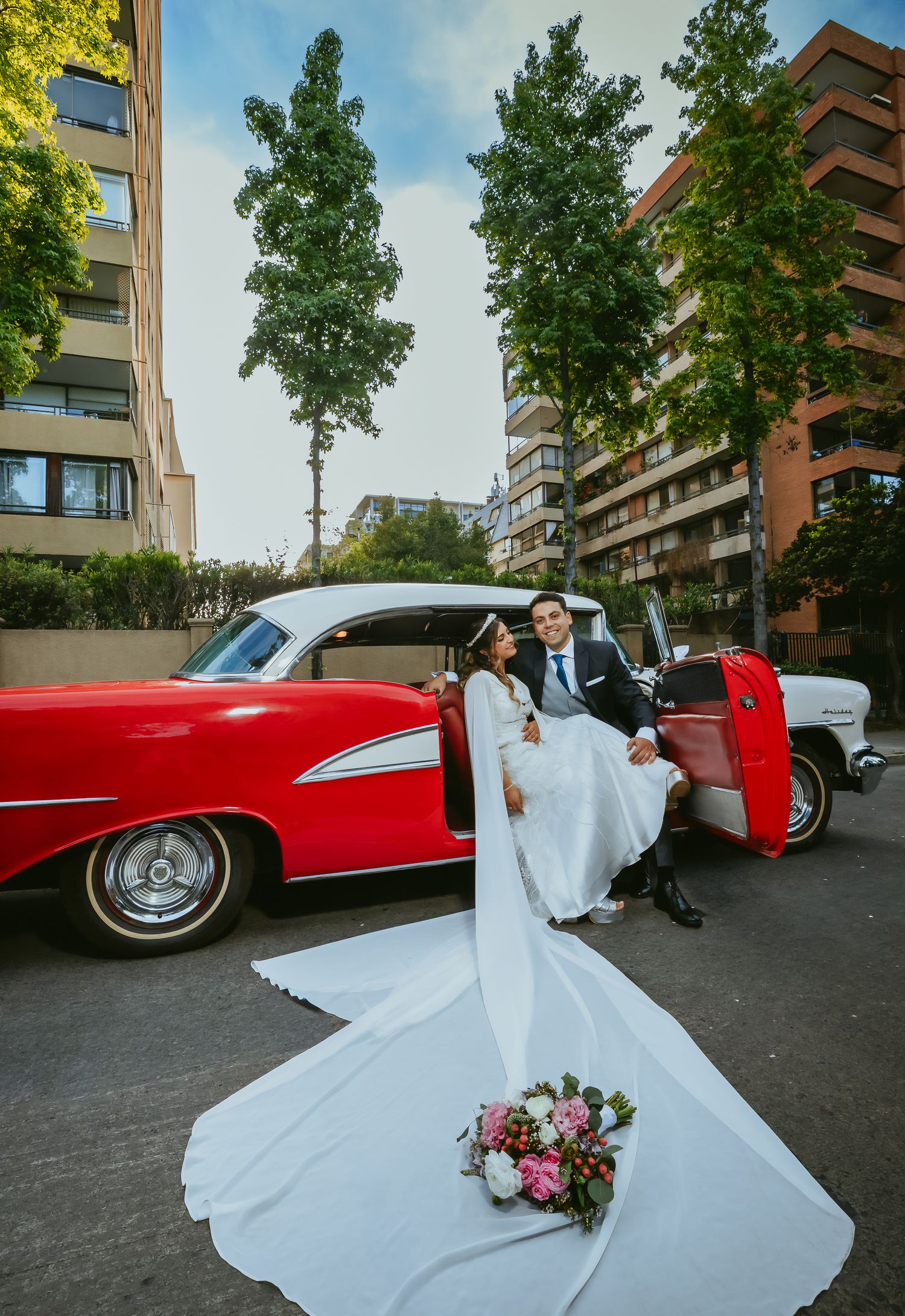 Bride in flowing white gown sitting with groom on red vintage classic car door on a sunny urban street.