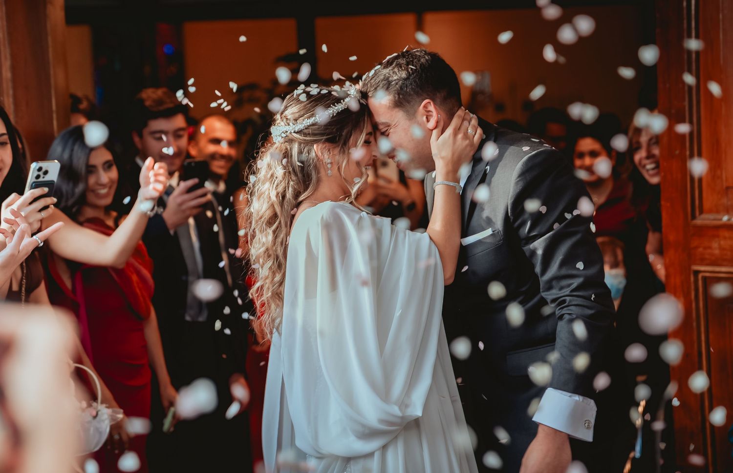 Bride and groom share a tender kiss surrounded by falling confetti and cheering wedding guests.