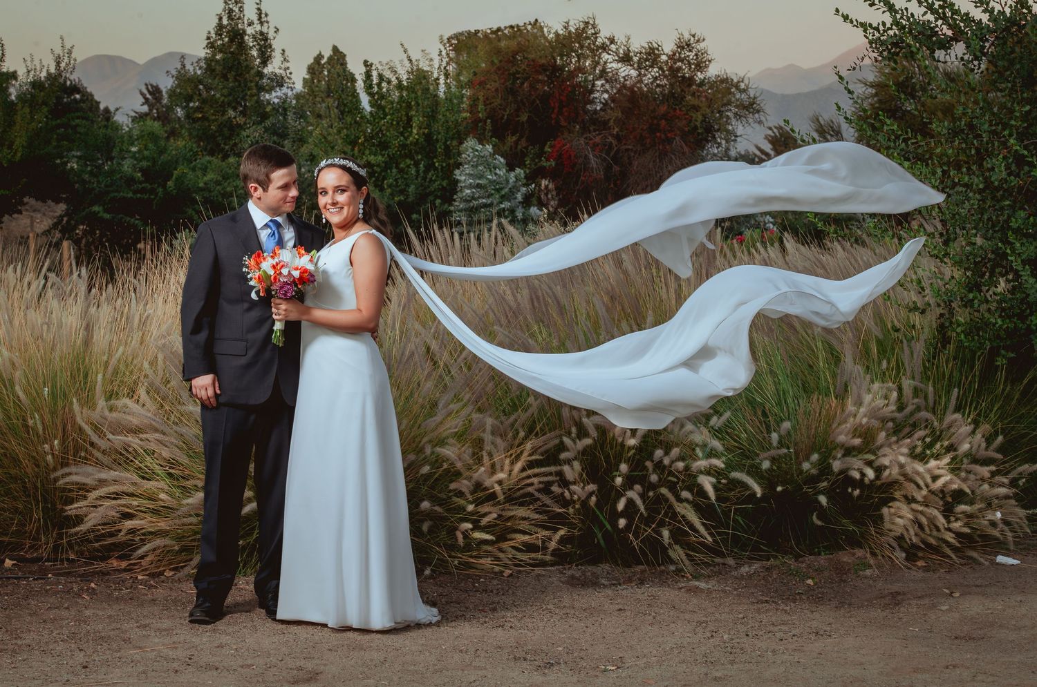 Bride's veil dramatically flowing in the wind as couple poses in an outdoor garden setting.