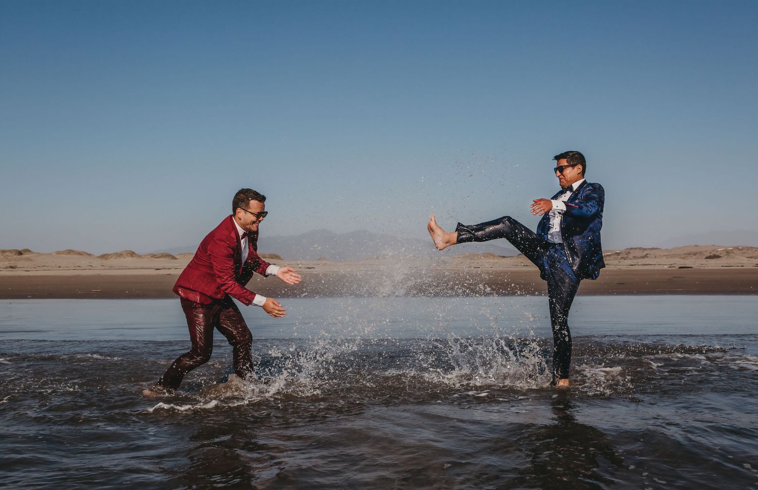 Two grooms playfully splash water at each other in a shallow beach setting under blue skies.