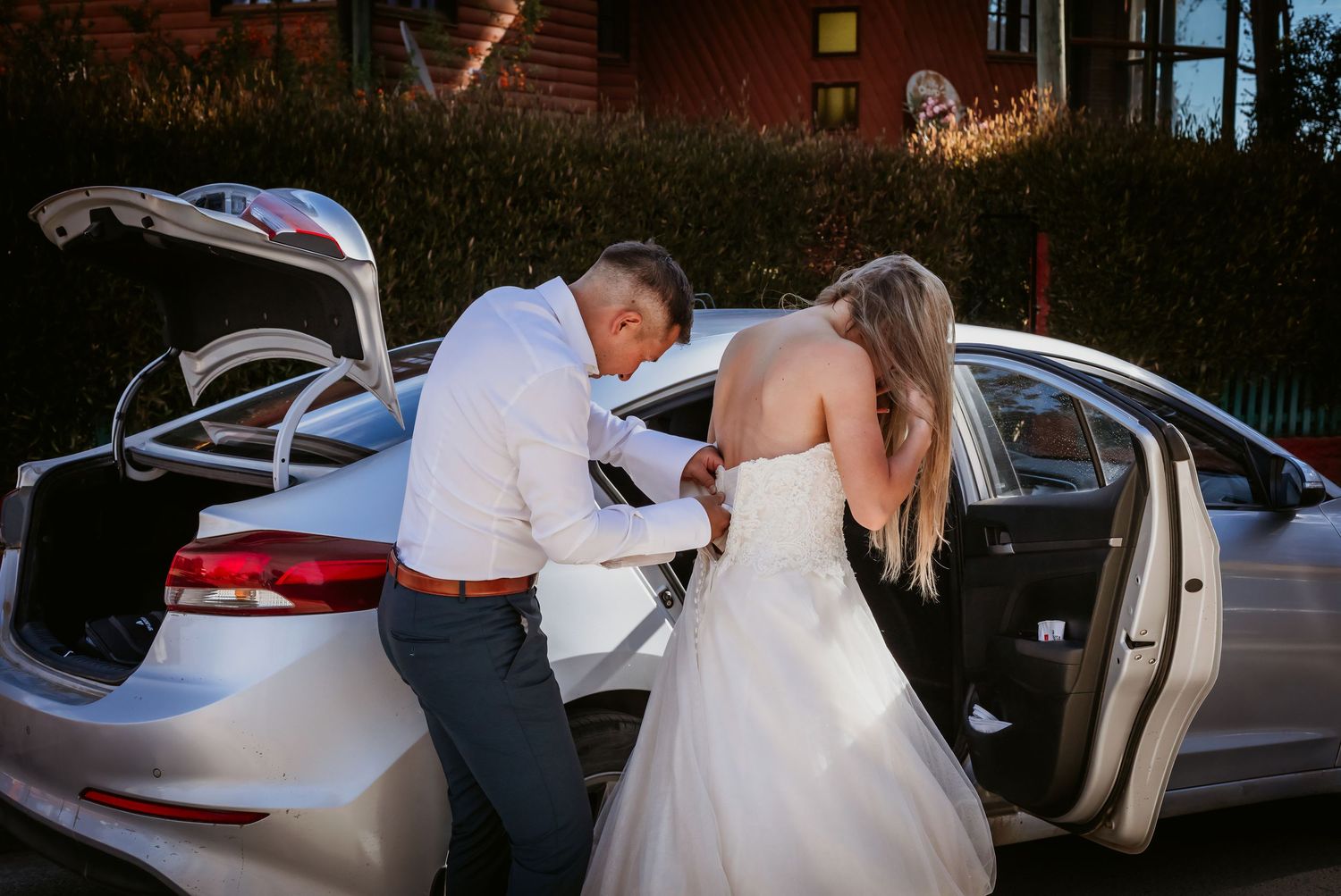 Groom helping bride with her wedding dress beside a white car.