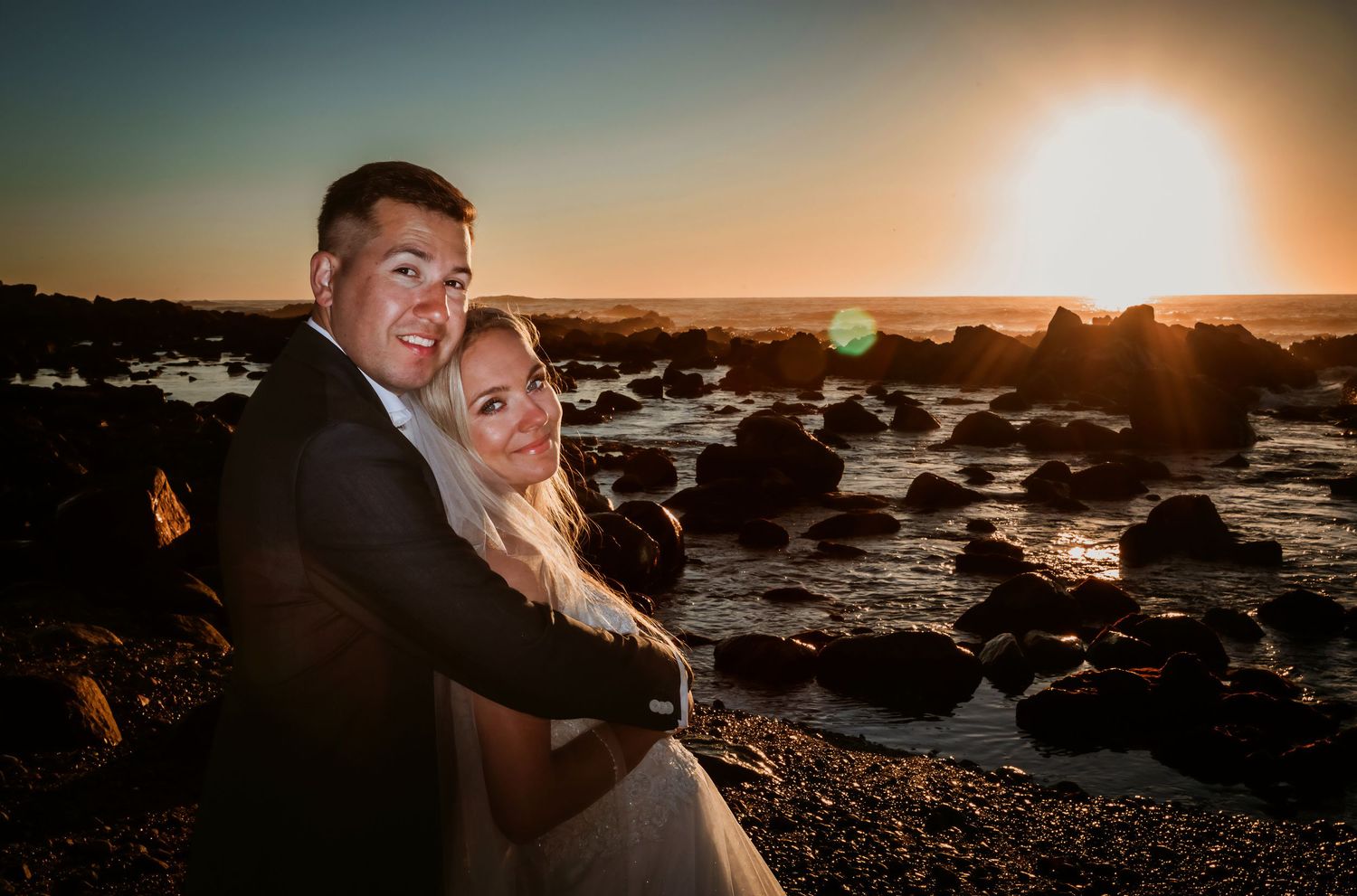 Smiling couple embracing on rocky beach at sunset, bride in strapless gown and groom in suit.