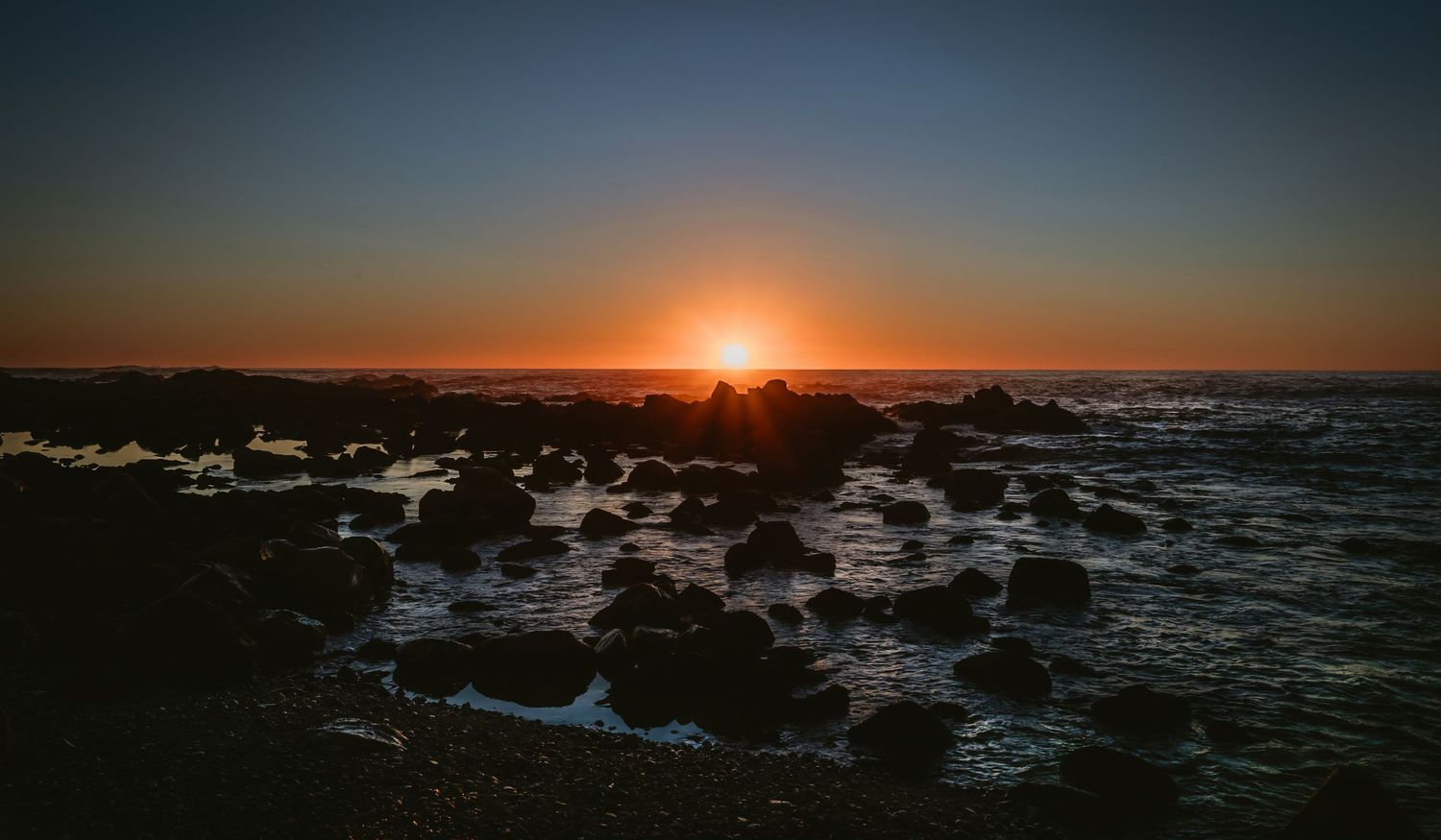 Dramatic coastal sunset with rocky shoreline silhouettes and glowing orange horizon over calm sea.