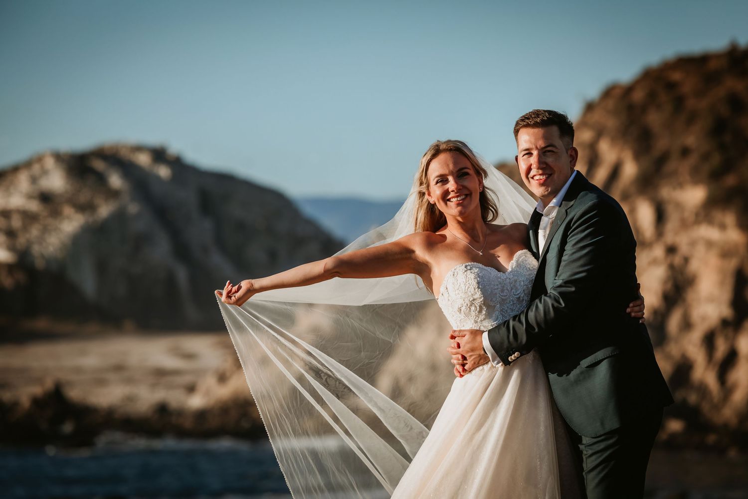 Happy couple embracing on rocky coastal cliffs, bride's veil flowing in the wind.