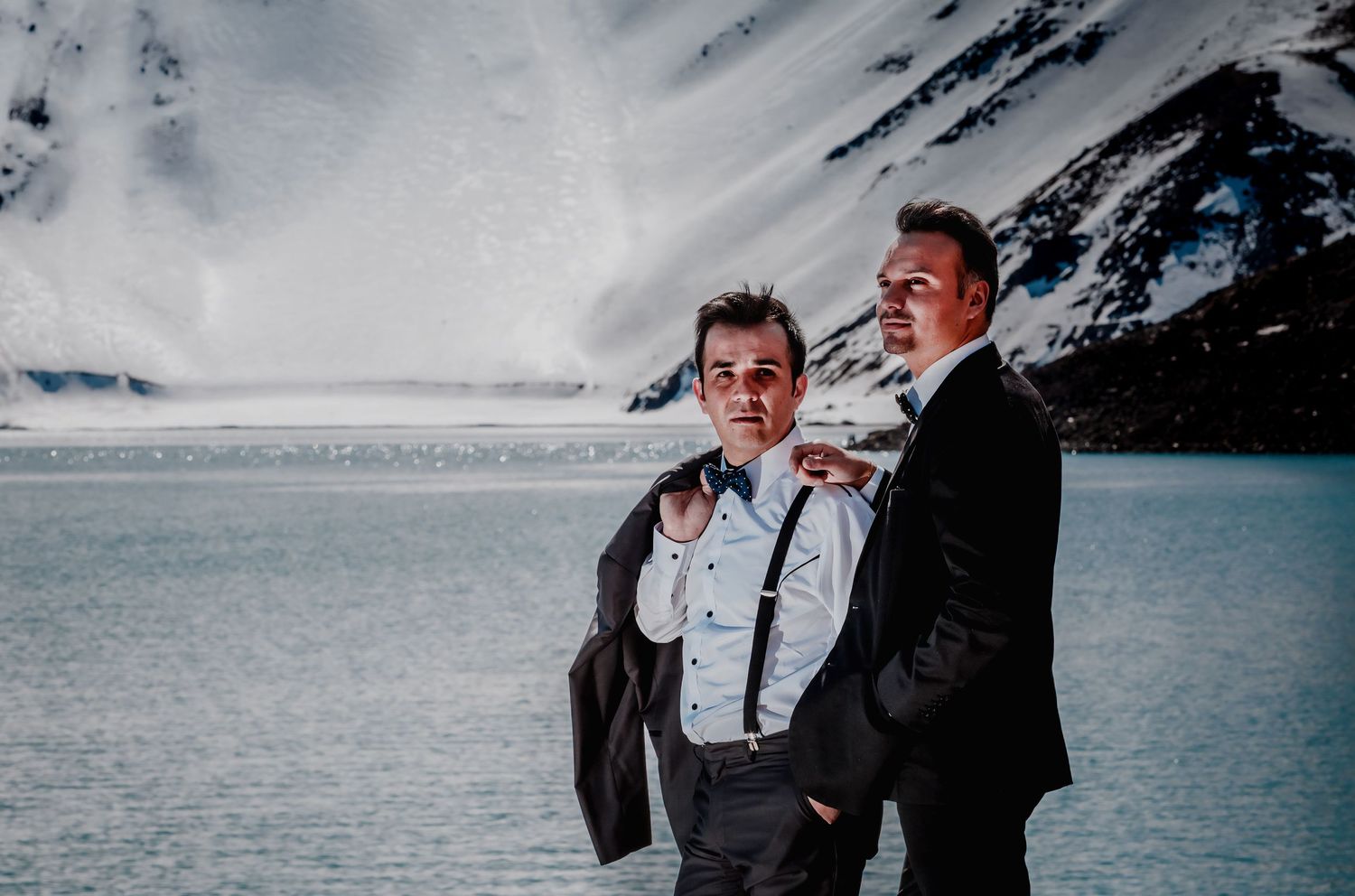 Two grooms pose in formal suits by a glacial lake with snow-capped mountains backdrop.