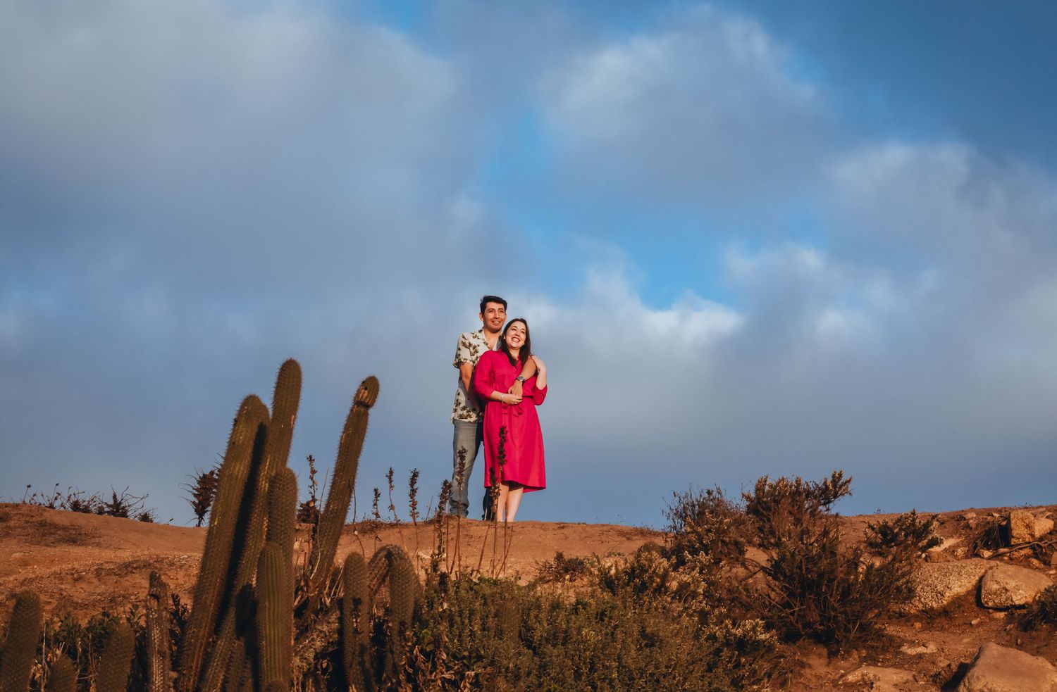 Couple posing on desert rocks with cacti and blue cloudy sky, woman in red dress, man in casual attire.