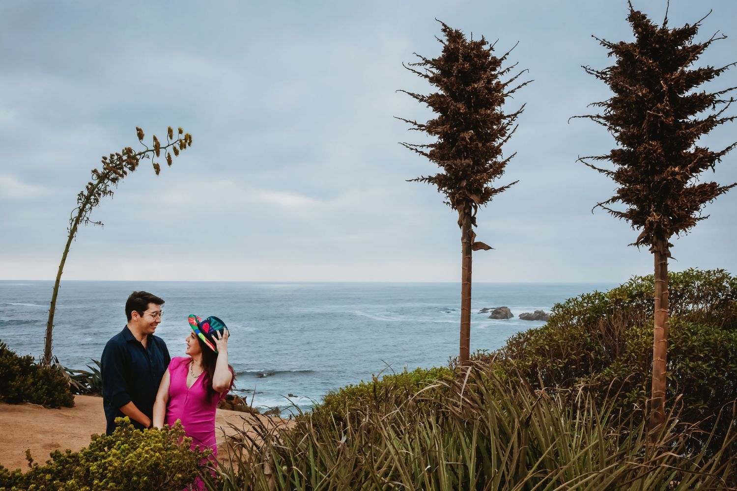 Couple standing together near coastal bluff with tall agave plants and ocean view on cloudy day.