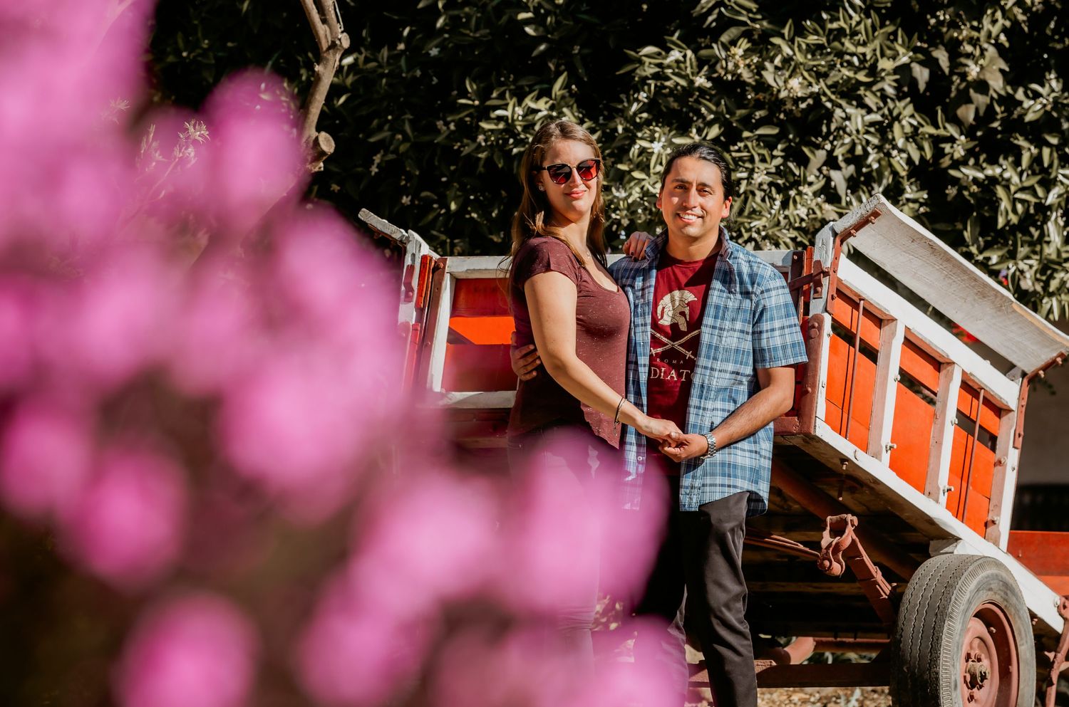 Couple smiling together beside a red farm trailer, framed by pink blossoms in the foreground.