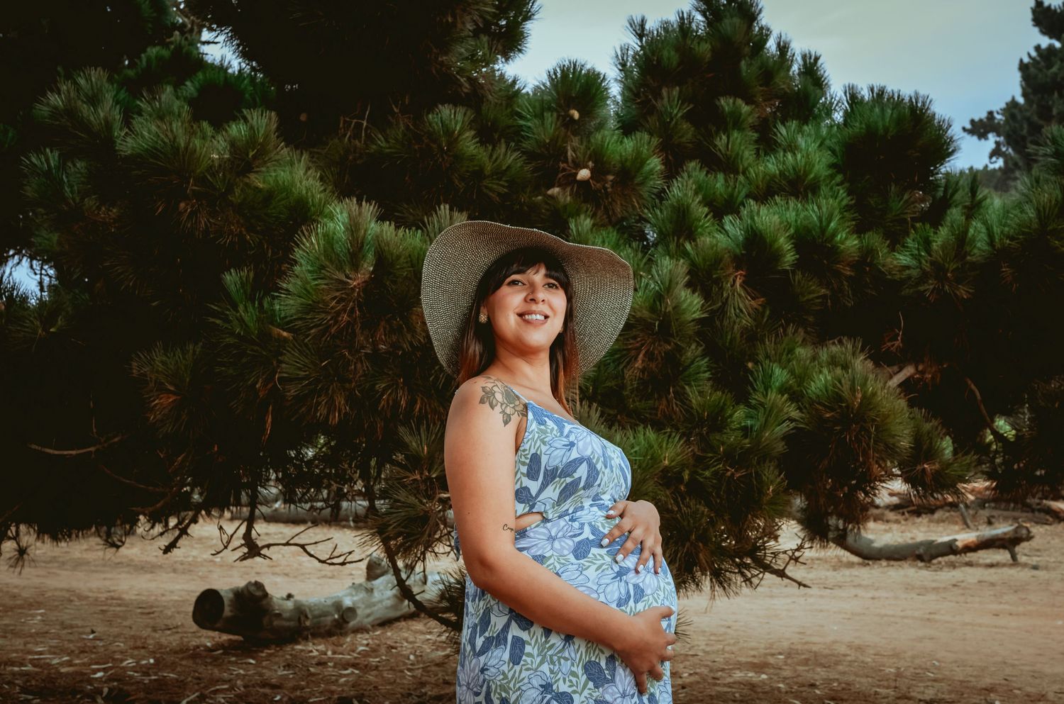 Smiling pregnant woman in blue floral dress and hat standing near pine trees.
