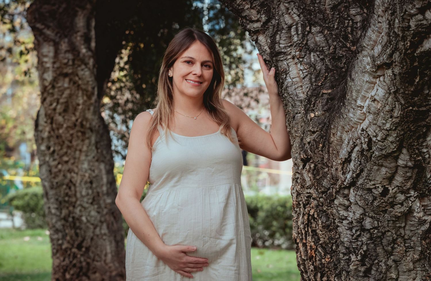Happy pregnant woman in white dress standing between tree trunks in a green park.