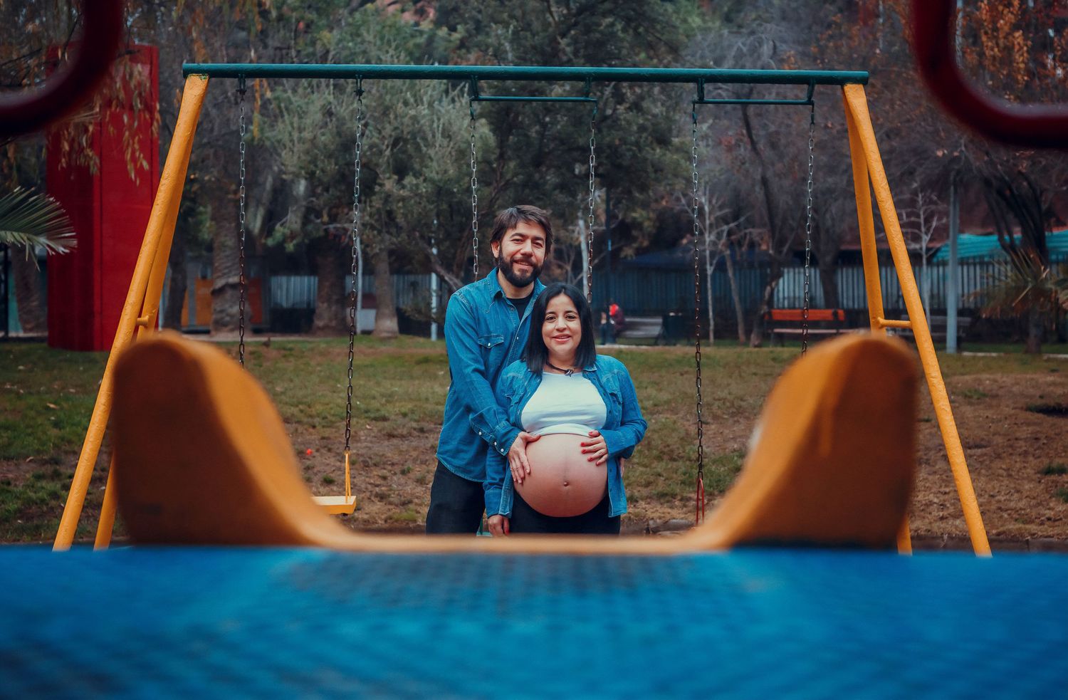 Couple posing at playground swing set, woman cradling pregnant belly, man standing behind her outdoors.