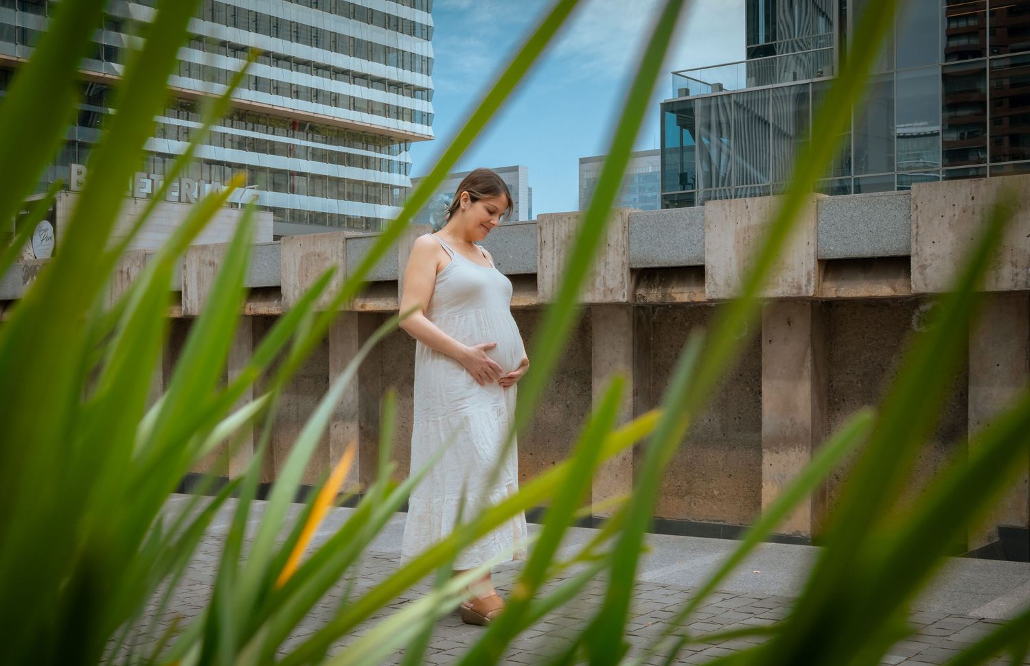 Pregnant woman in a white dress cradling her baby bump outdoors near modern urban architecture and greenery.