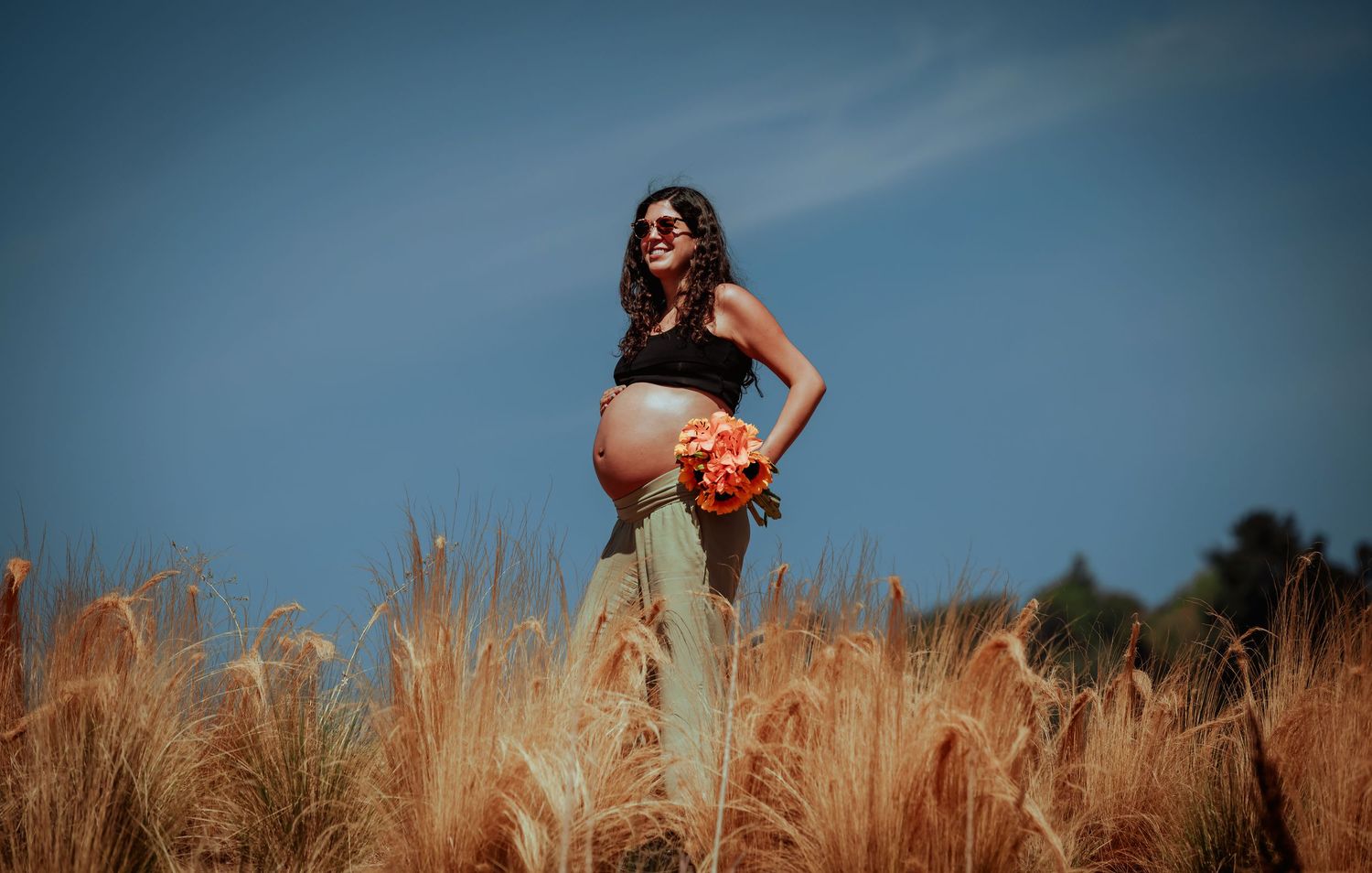 Pregnant woman in sunglasses standing in a golden wheat field holding an orange bouquet under blue sky.