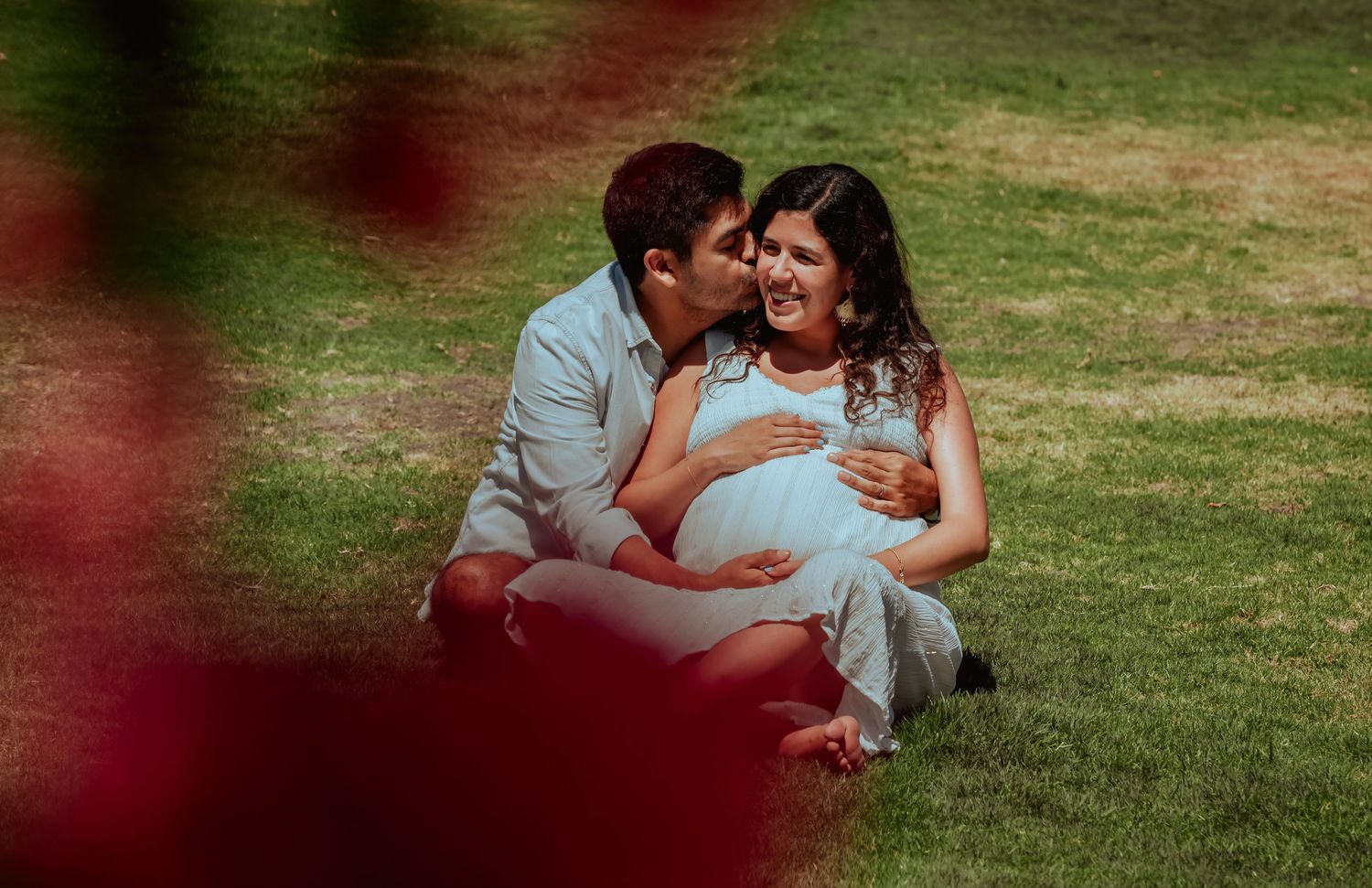 Man kissing pregnant woman's cheek while holding her baby bump, sitting on grass in sunny park.