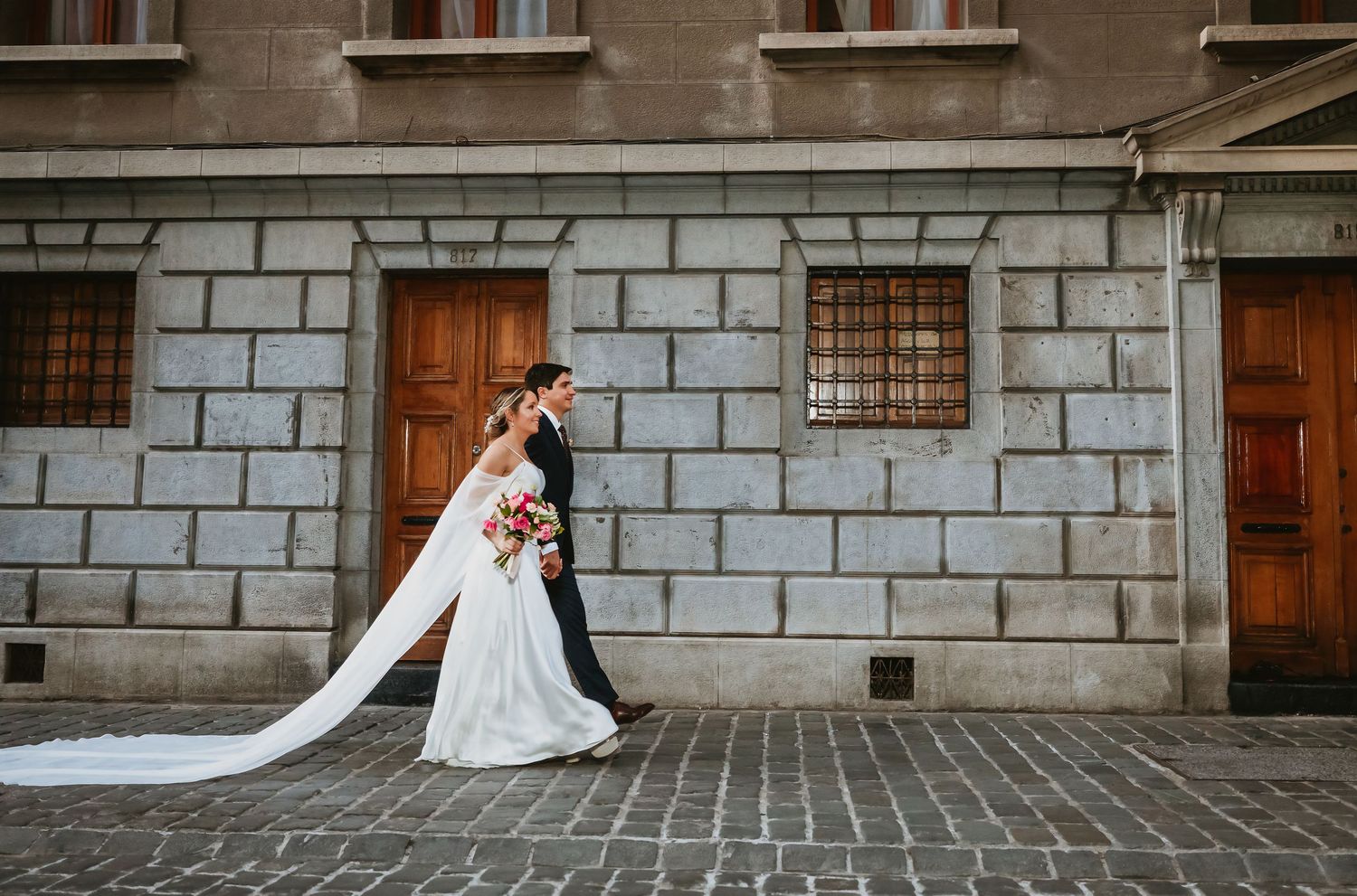 Bride and groom sharing a kiss on cobblestone street beside stone building, bride holding bouquet.