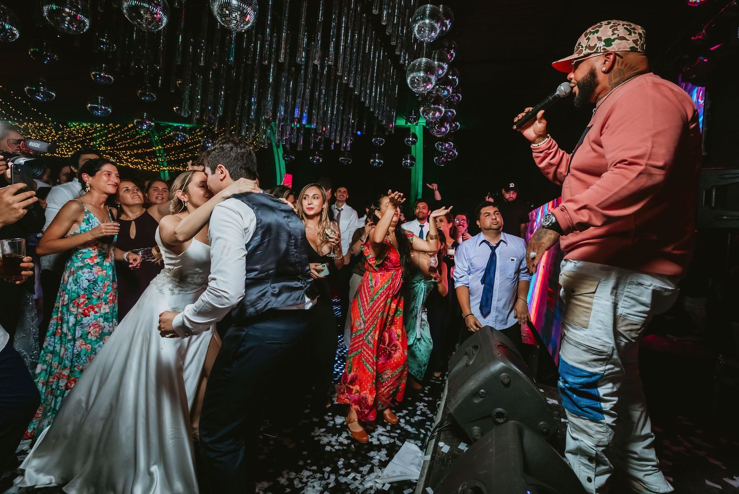 A bride and groom share a romantic kiss on the dance floor surrounded by celebrating guests at their wedding reception.