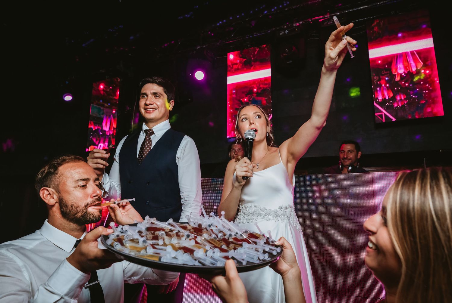 Bride holds microphone while groom and guests enjoy food at a lively wedding reception.
