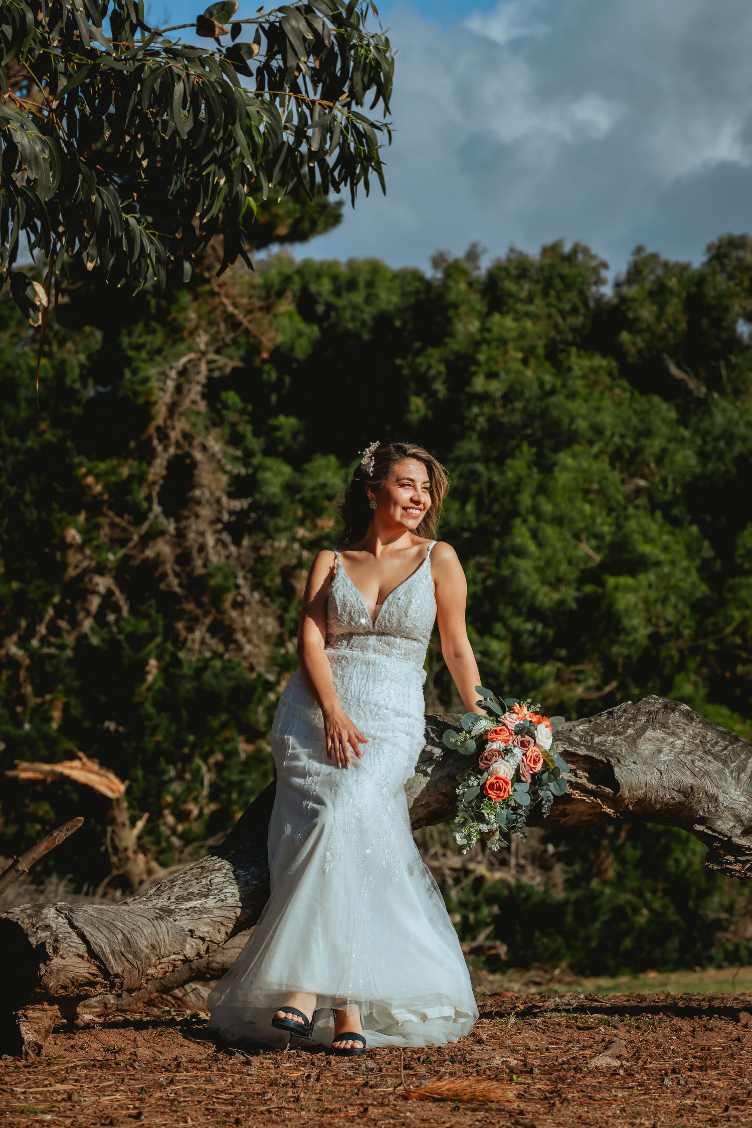 Smiling bride in a fitted lace wedding gown holds an orange and coral bouquet while sitting on a fallen log amid lush green trees.
