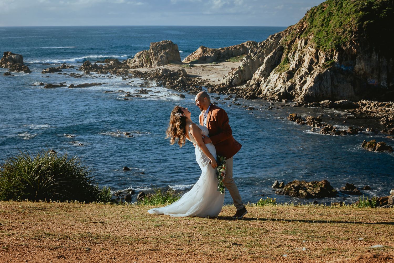 Bride in a white gown and groom in a rust jacket walk hand in hand on a grassy clifftop overlooking rocky coastline and blue ocean.