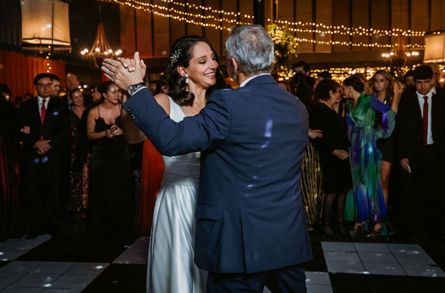 Bride and groom share first dance at elegant wedding reception with string lights and guests in background.