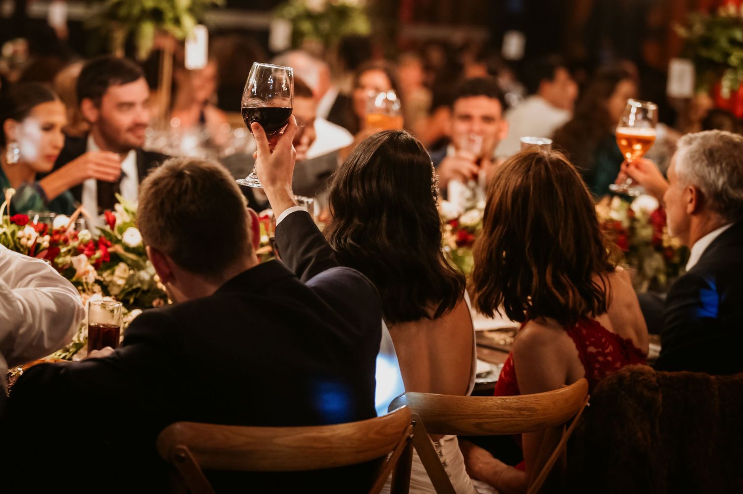 Elegant wedding reception guests raising glasses of red wine in a toast at a candlelit dinner table.
