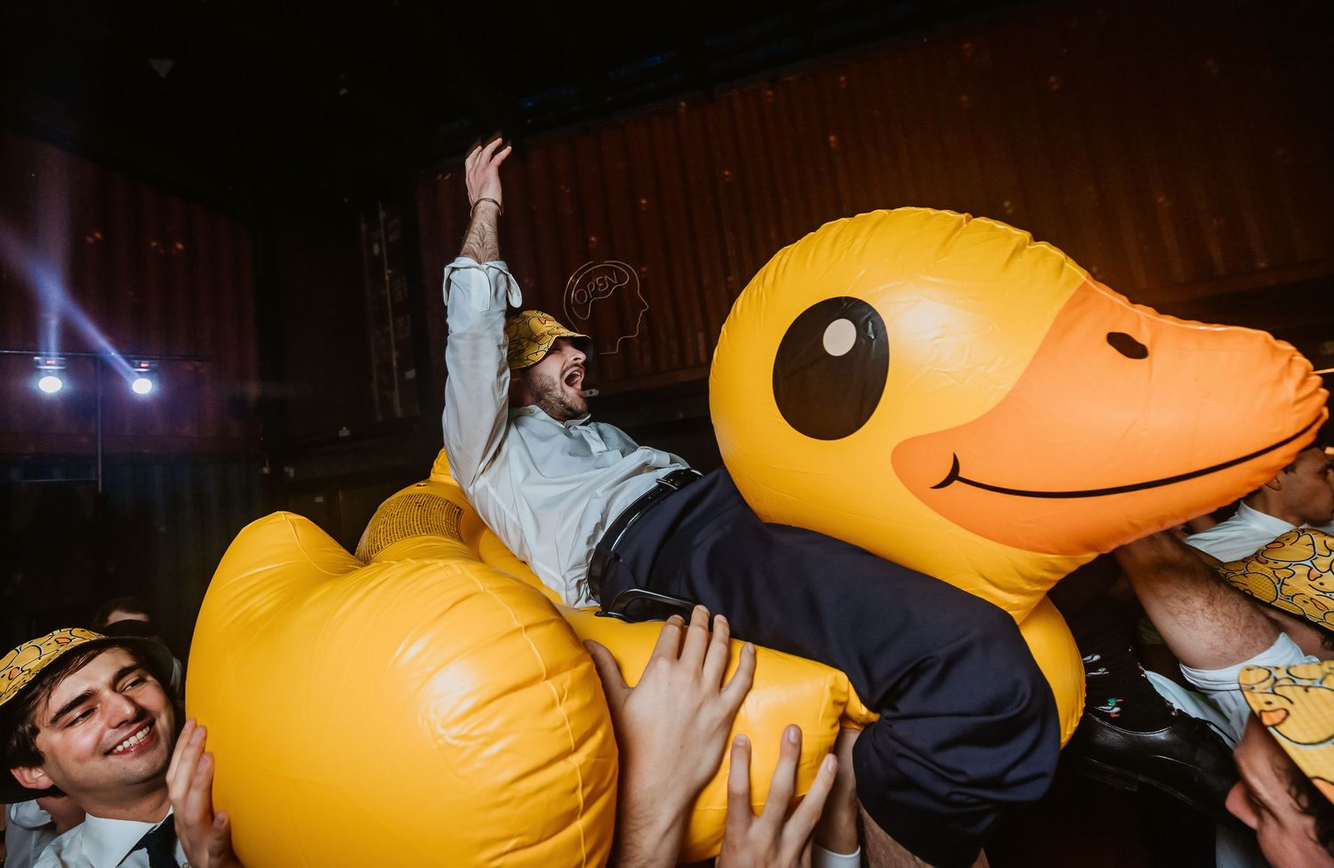 Man riding giant inflatable yellow rubber duck while crowd surfing at party.