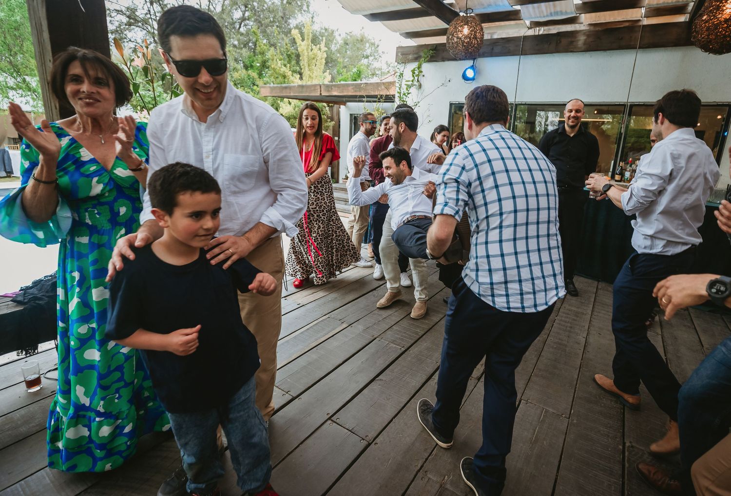 Adults and children dancing joyfully at an outdoor celebration on a wooden deck surrounded by guests.