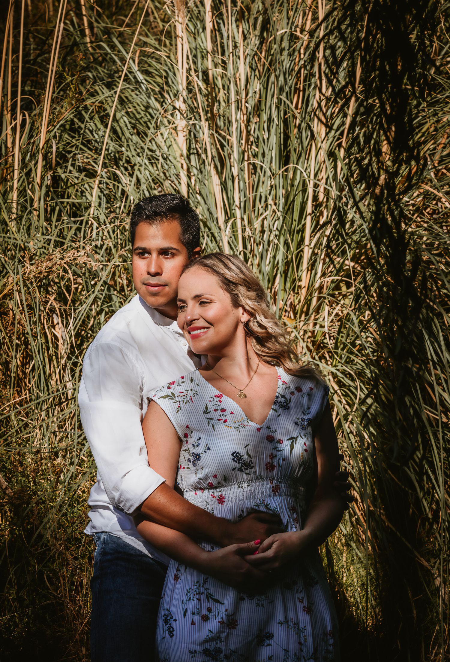 Couple embracing in front of tall golden grass, woman in floral dress smiling warmly during outdoor engagement photo session.