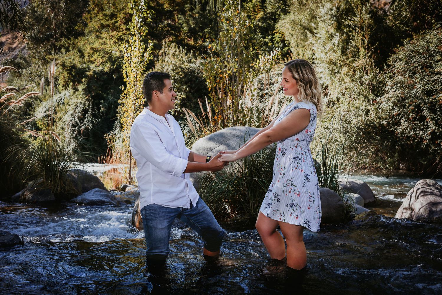 A man kneels on one knee proposing to a woman in a flowing floral dress beside a rocky stream outdoors.