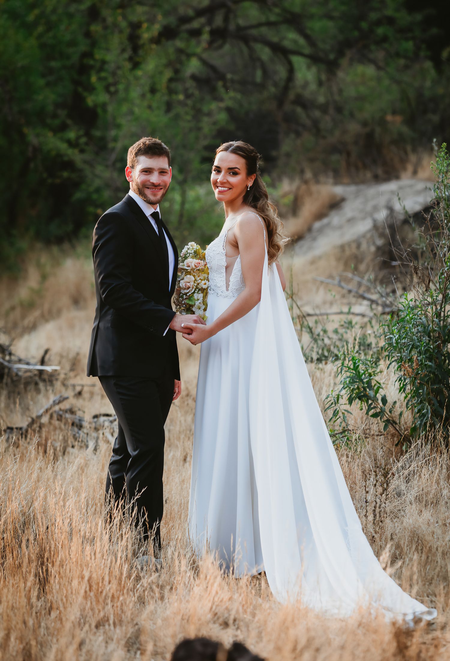 Smiling bride in white gown holding bouquet stands with groom in black tuxedo in a scenic outdoor field.