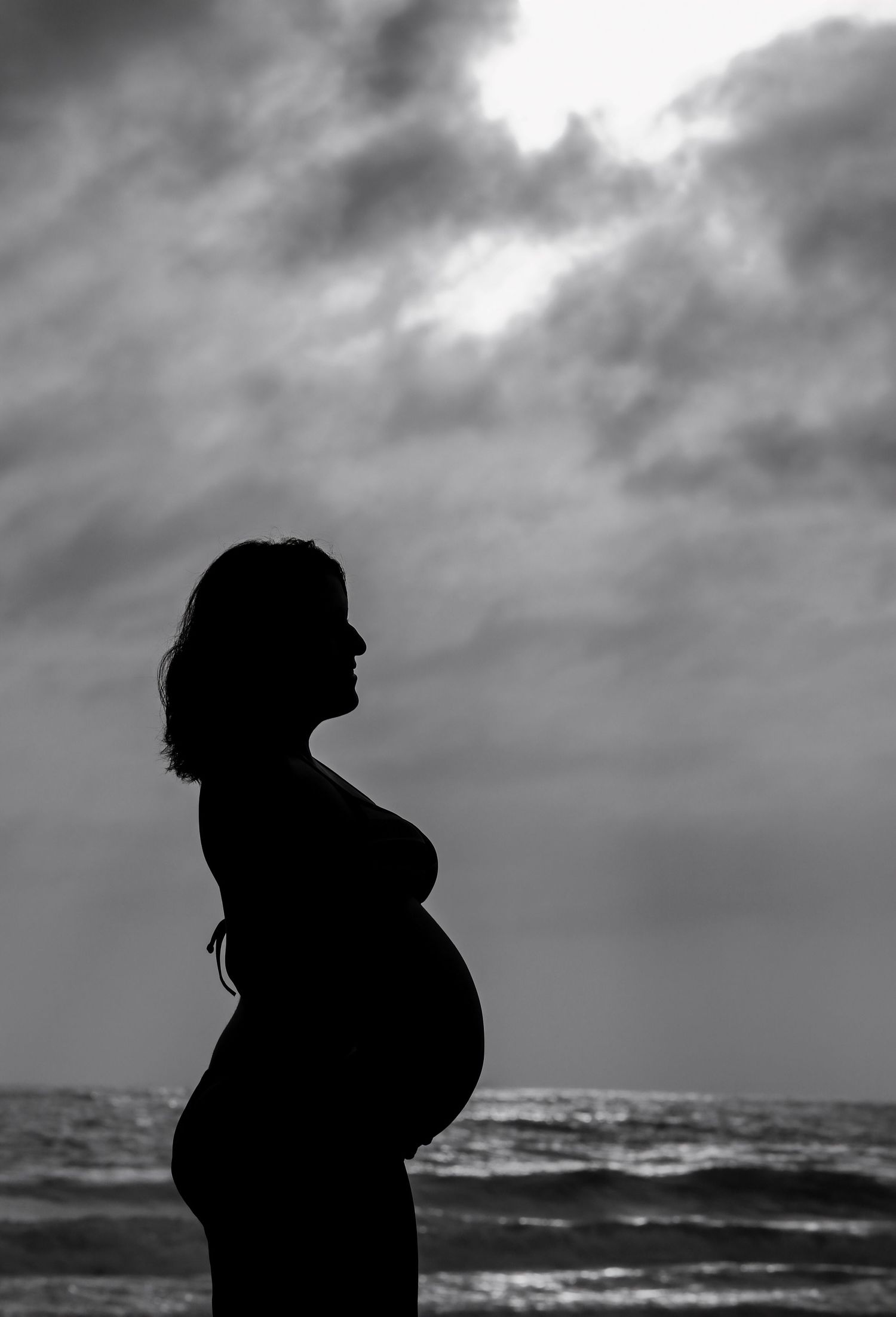 Silhouette of a pregnant woman standing by the ocean against a dramatic cloudy sky in black and white.