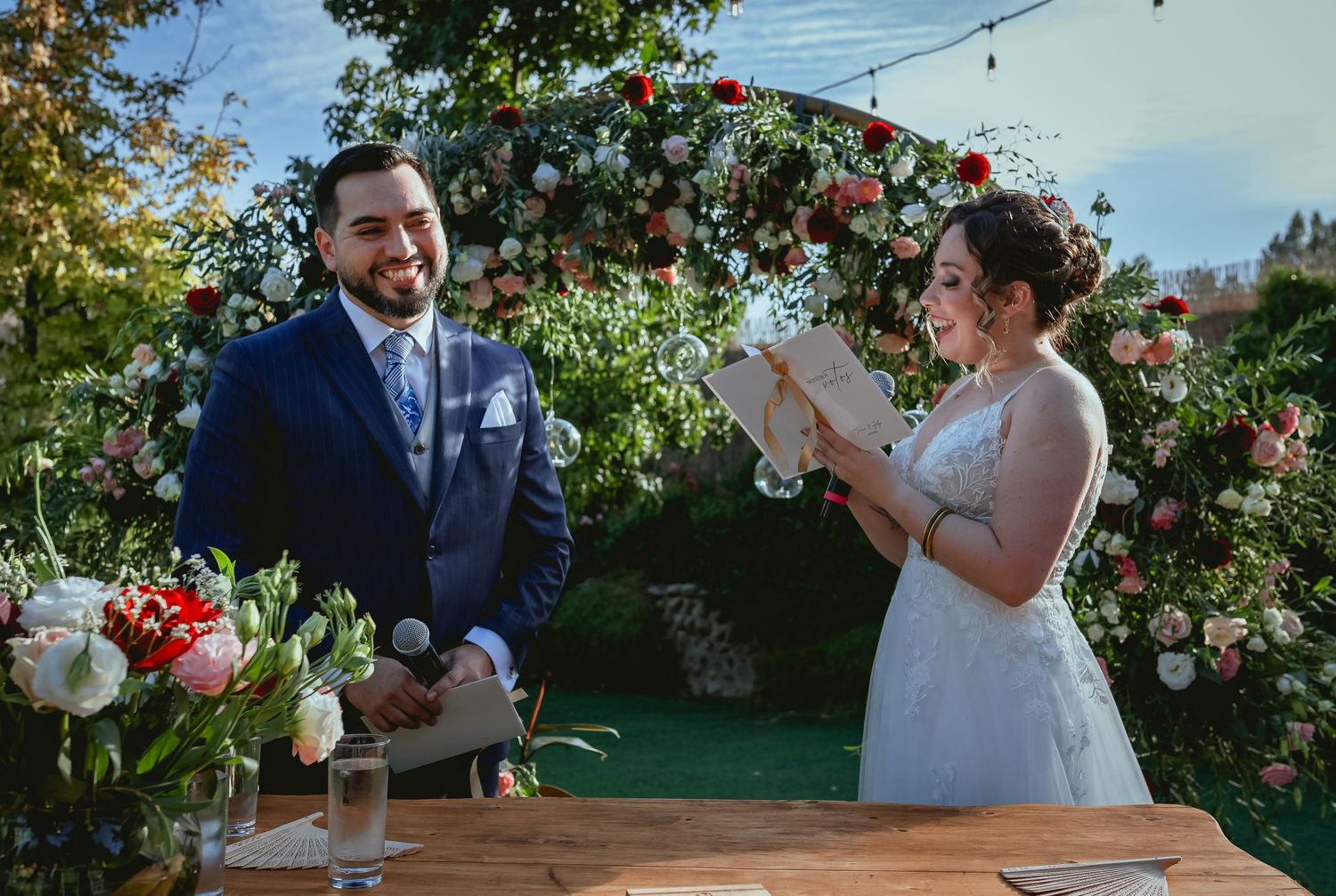 Bride reads heartfelt vows to smiling groom at outdoor wedding ceremony beneath floral arch.