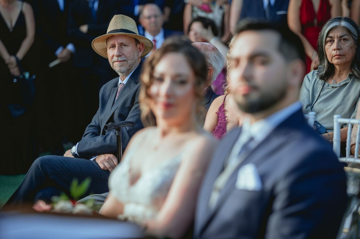 Wedding guest in straw hat watches ceremony as bride and groom appear blurred in foreground outdoors.
