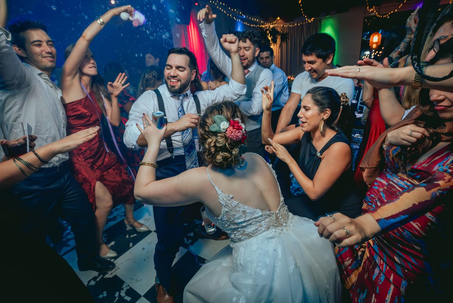 Bride dancing joyfully with wedding guests under colorful lights at reception.