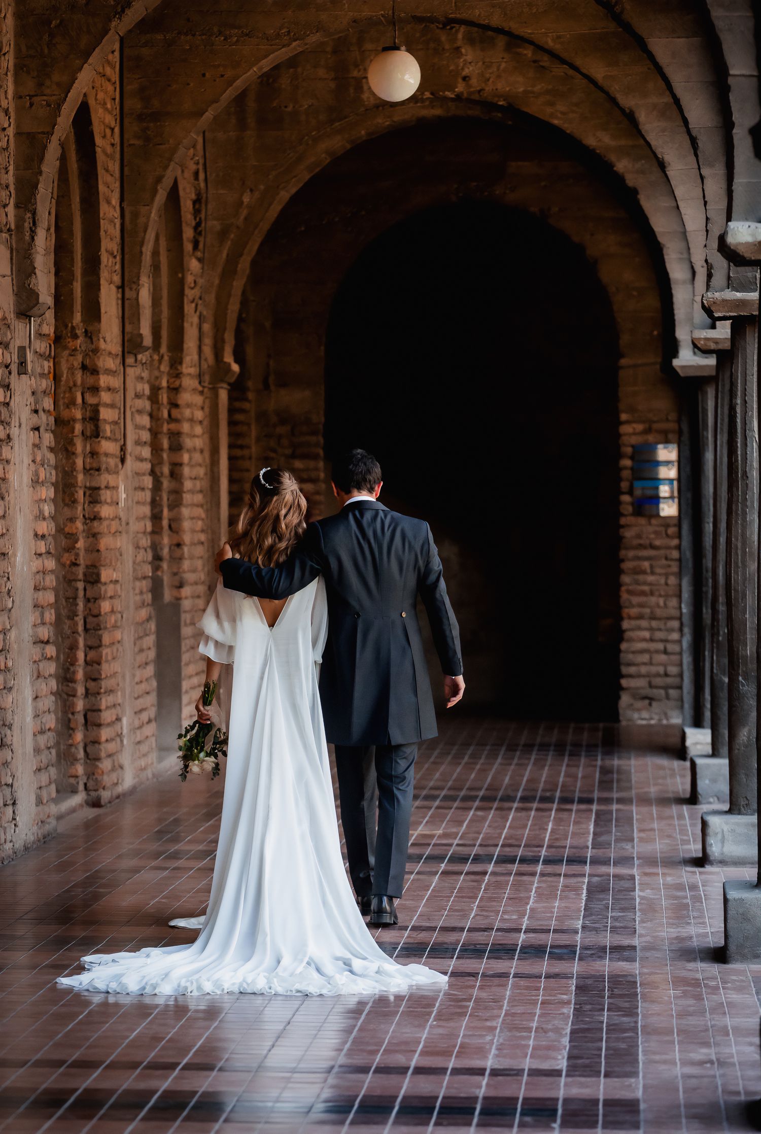 Bride and groom walking hand in hand through a brick arched corridor, viewed from behind.